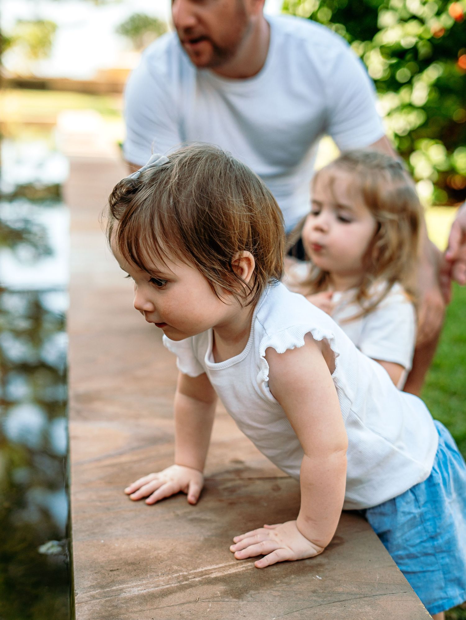 Two young girls in white tops curiously lean over a wooden ledge near a garden pond.