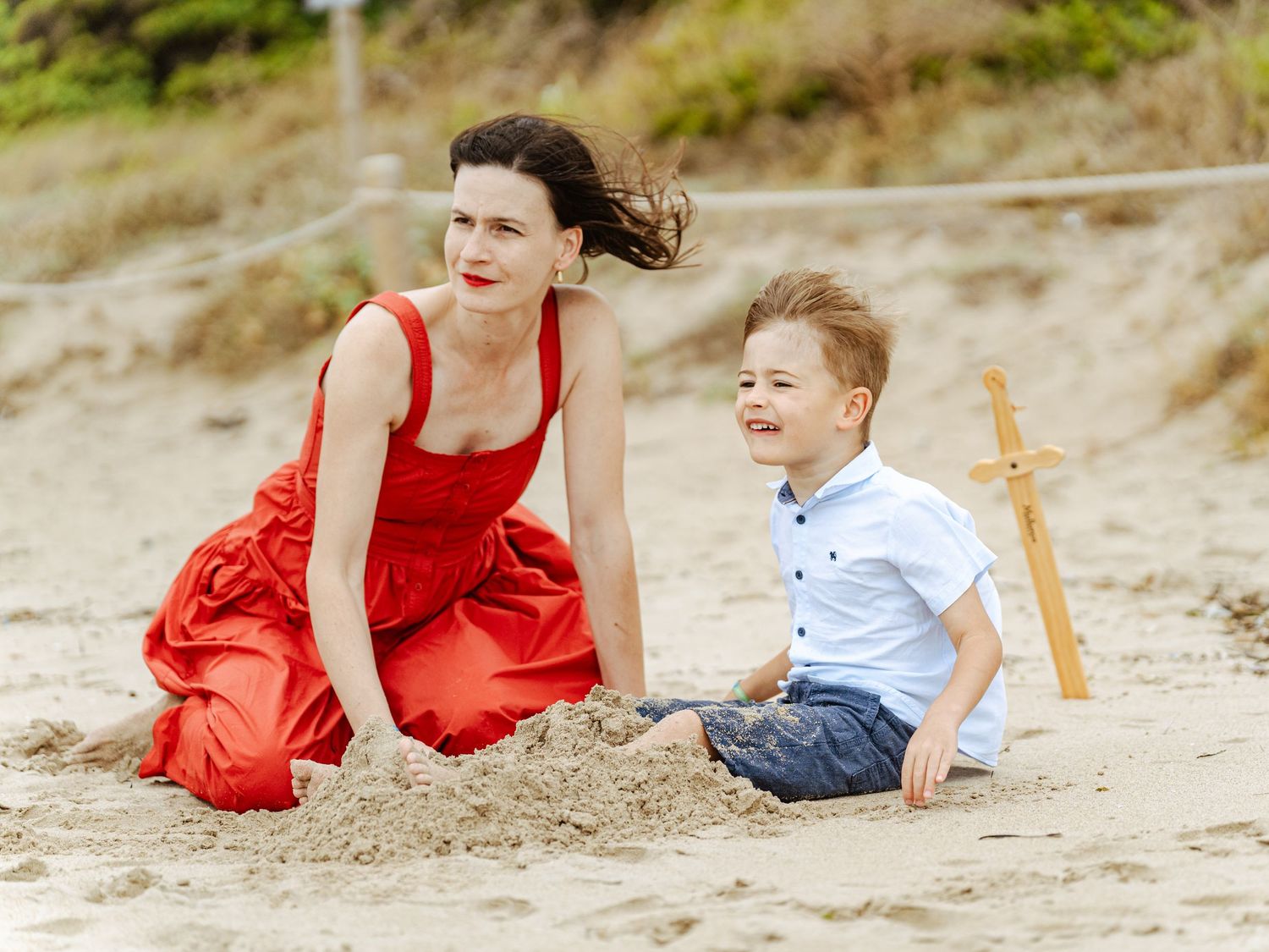 Mother in flowing red dress sits with laughing young son on sandy beach.