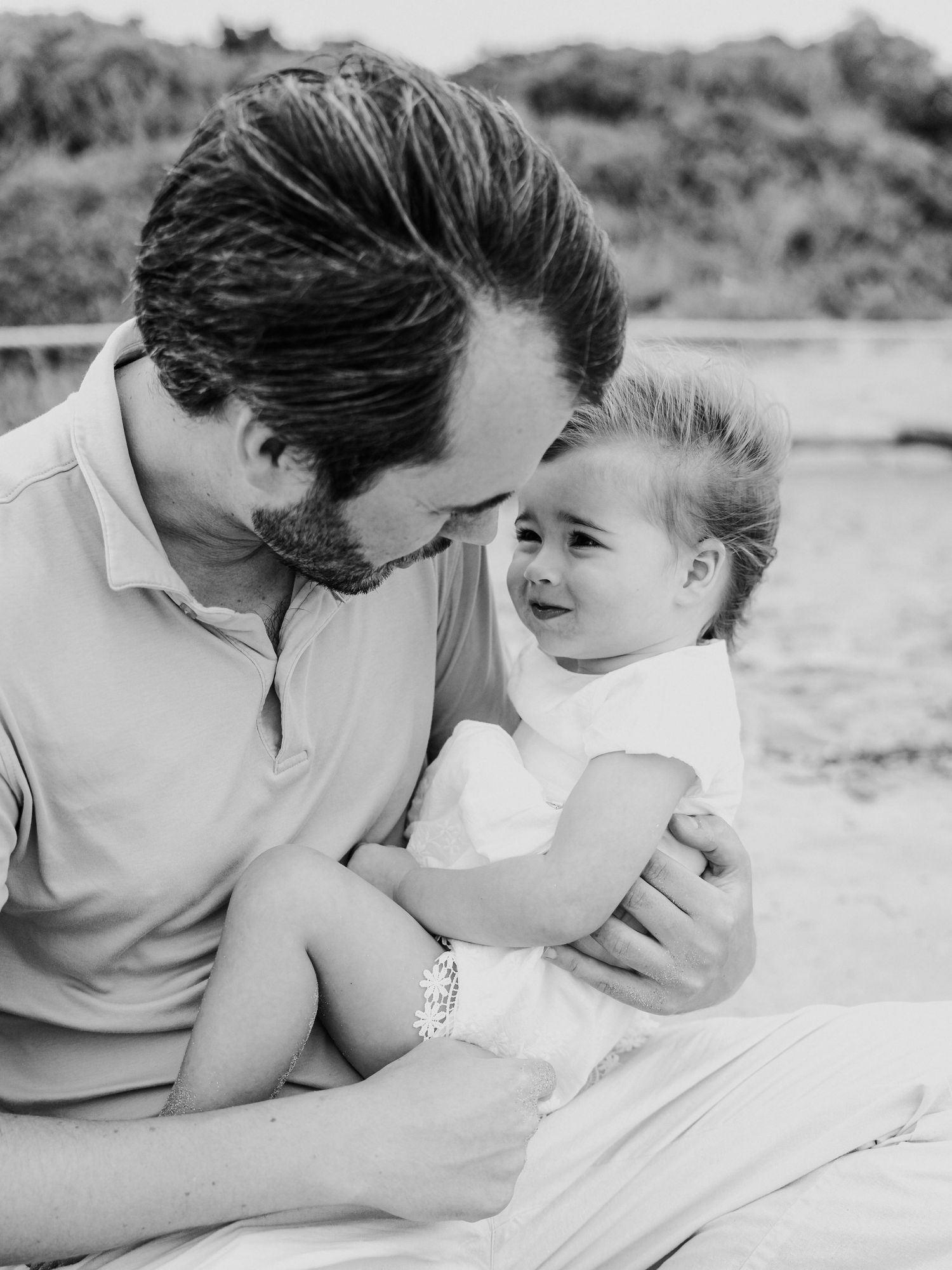 Black and white photo of bearded father holding laughing baby on sandy beach.