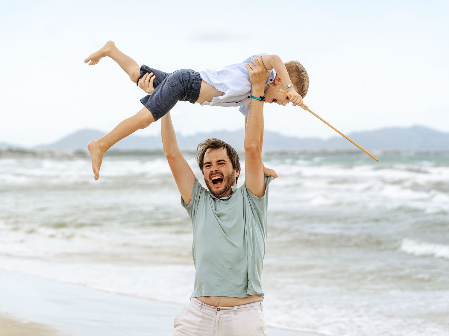Joyful father lifting child high above head on sunny beach with mountains.