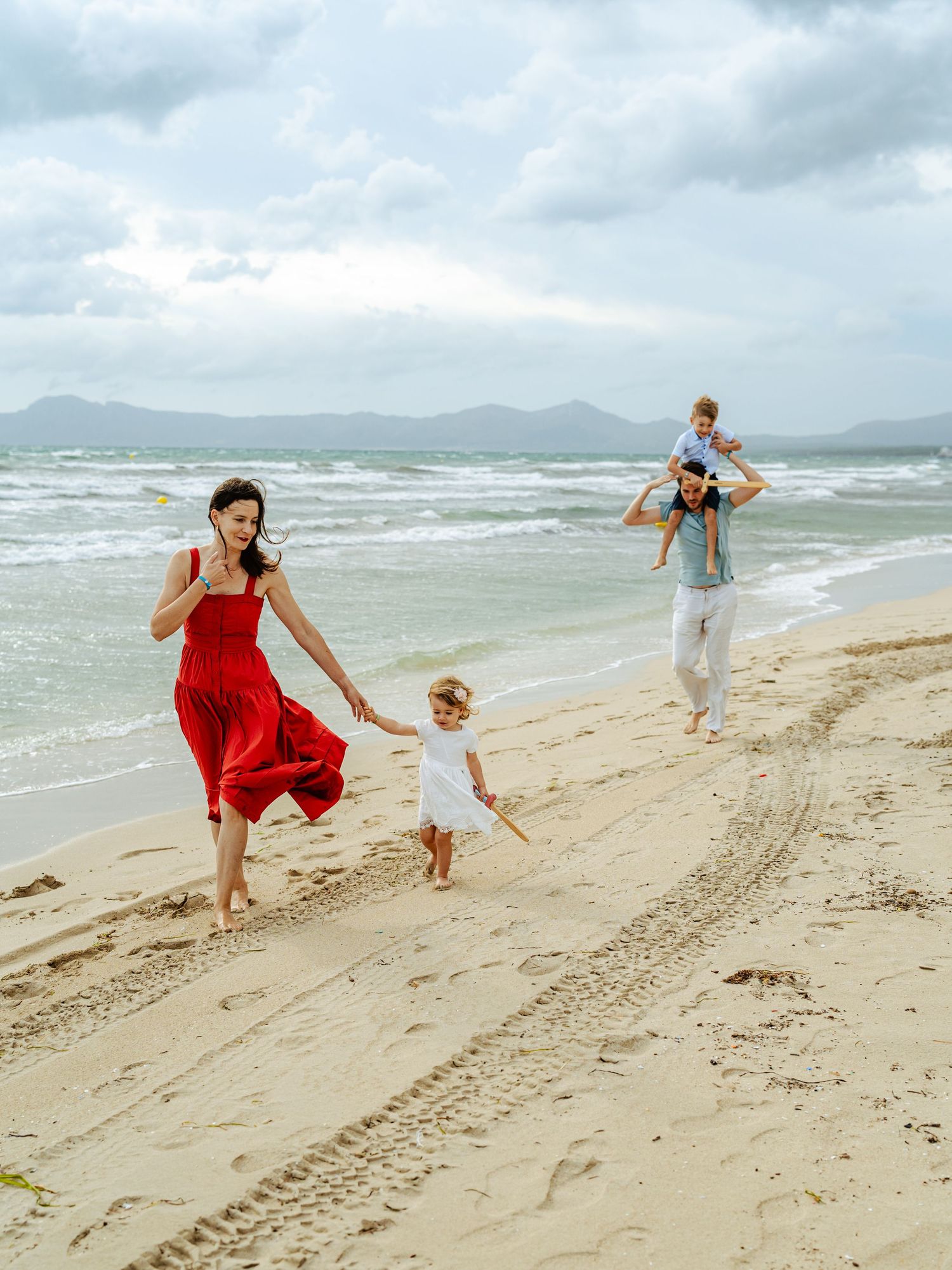 Family walking on windy beach, mother in red dress holds toddler while father carries son.