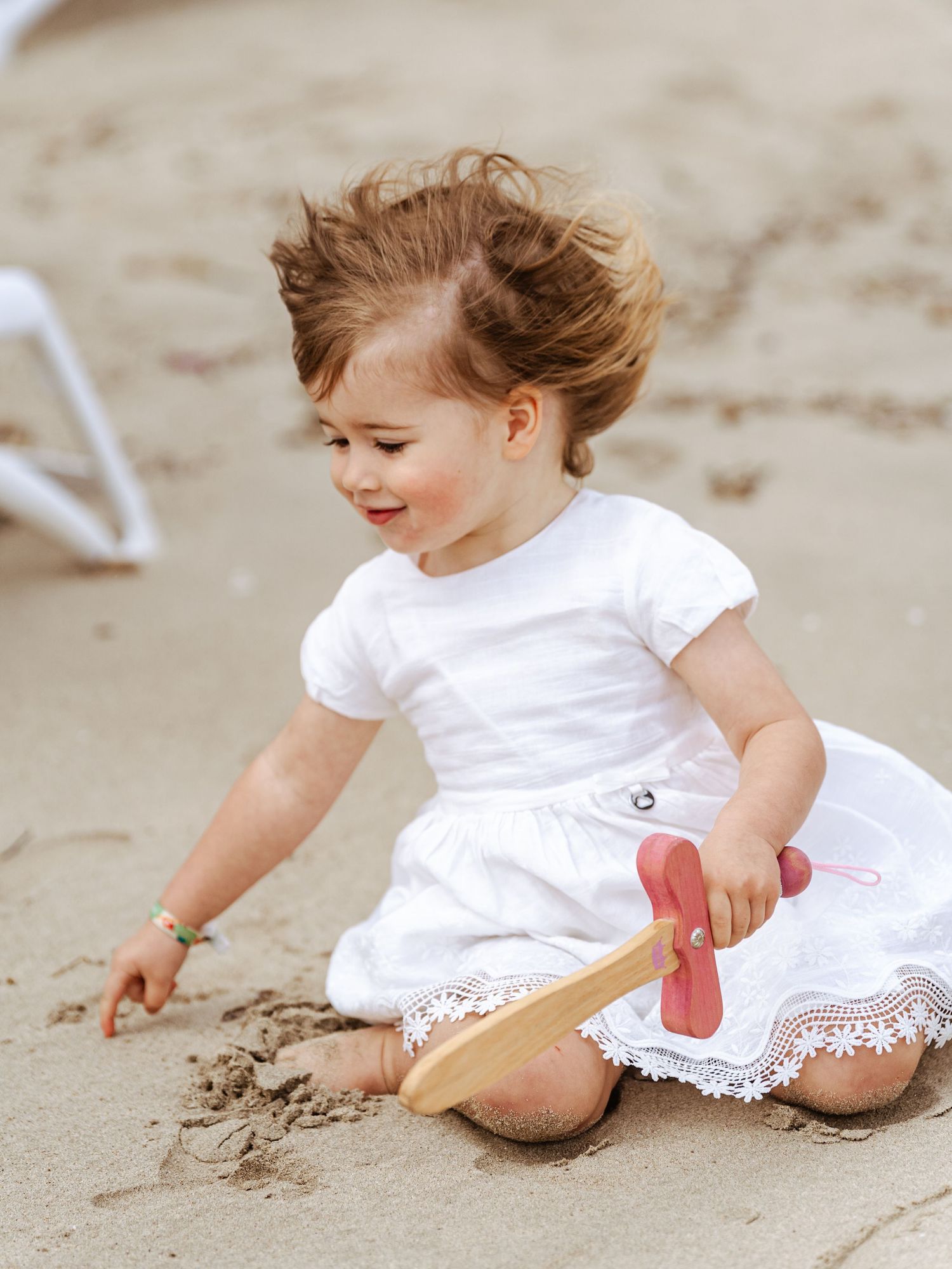 A cute toddler girl in a white lace dress plays in the sand with a wooden toy on the beach.