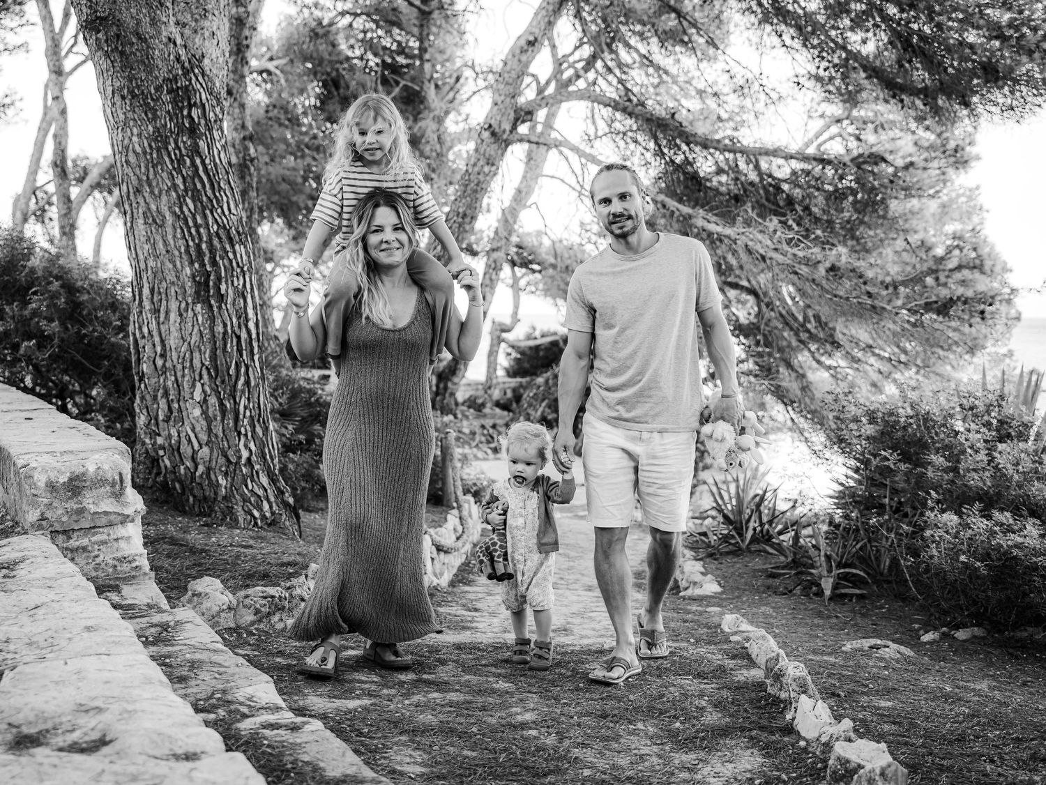 Black and white photo of joyful family walking along garden path, mother with child on shoulders.