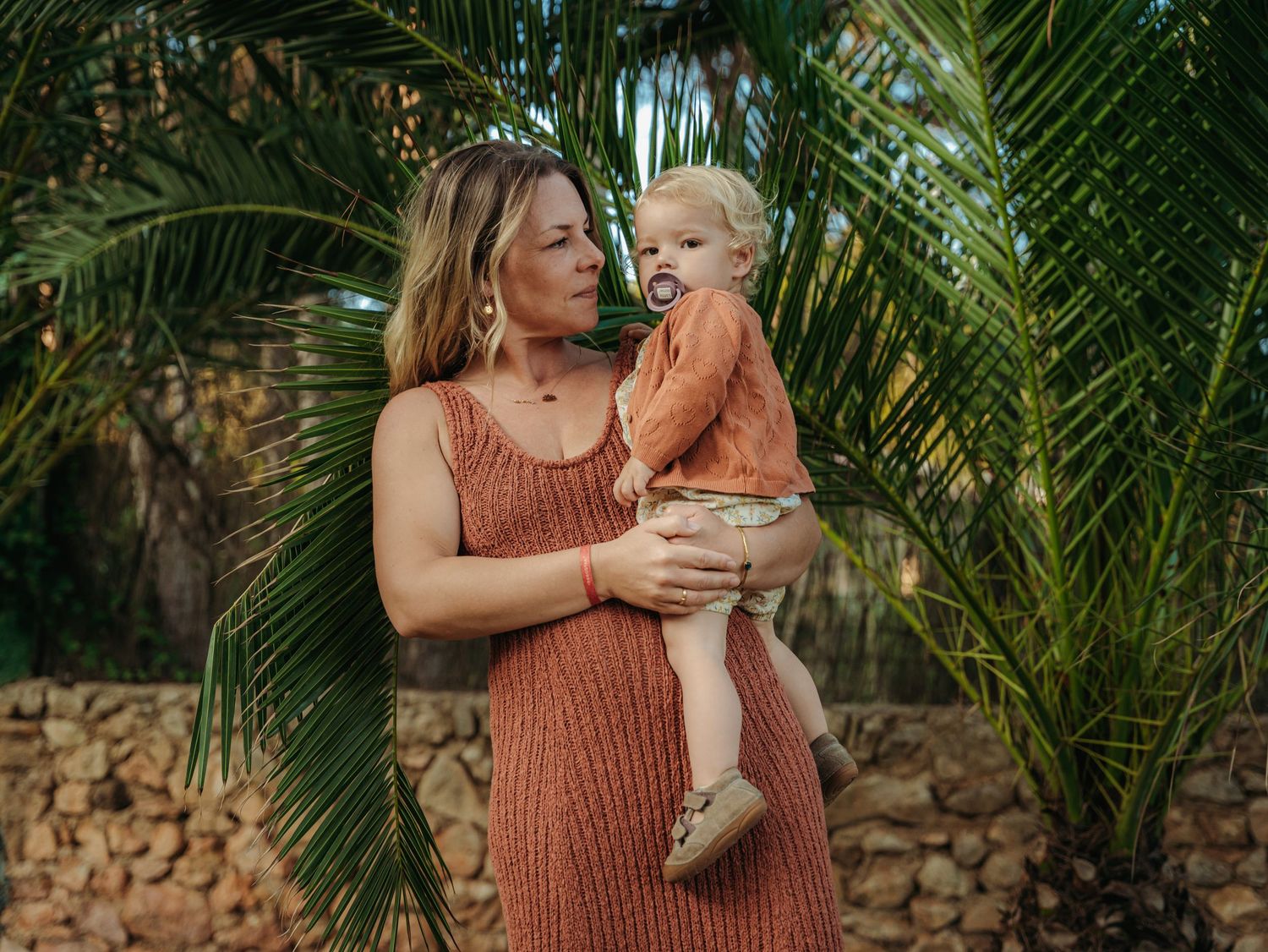 Mother in terracotta dress holding toddler son with pacifier beside lush palm trees.