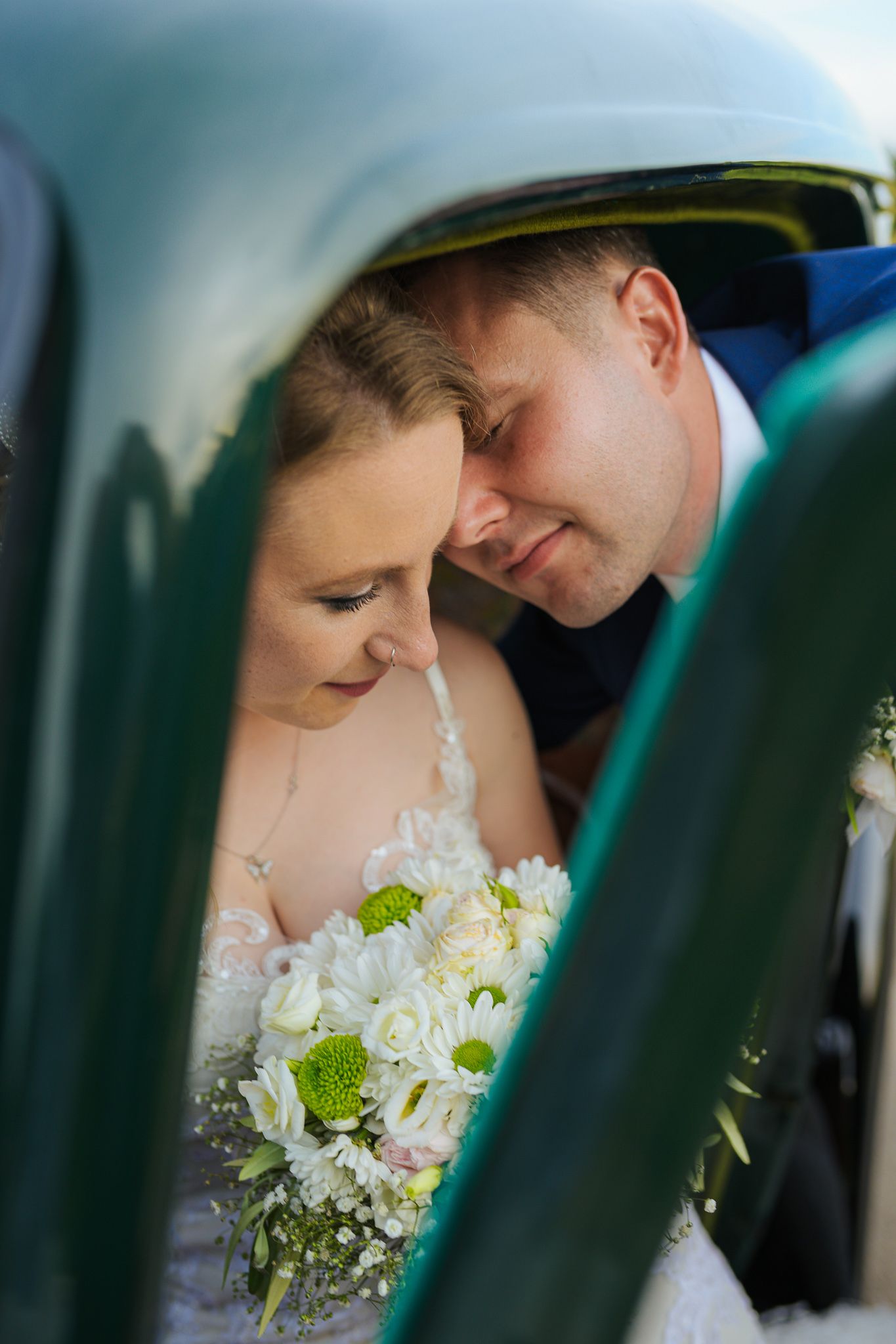 Newlyweds sharing an intimate moment together inside a vintage teal car with bridal bouquet.