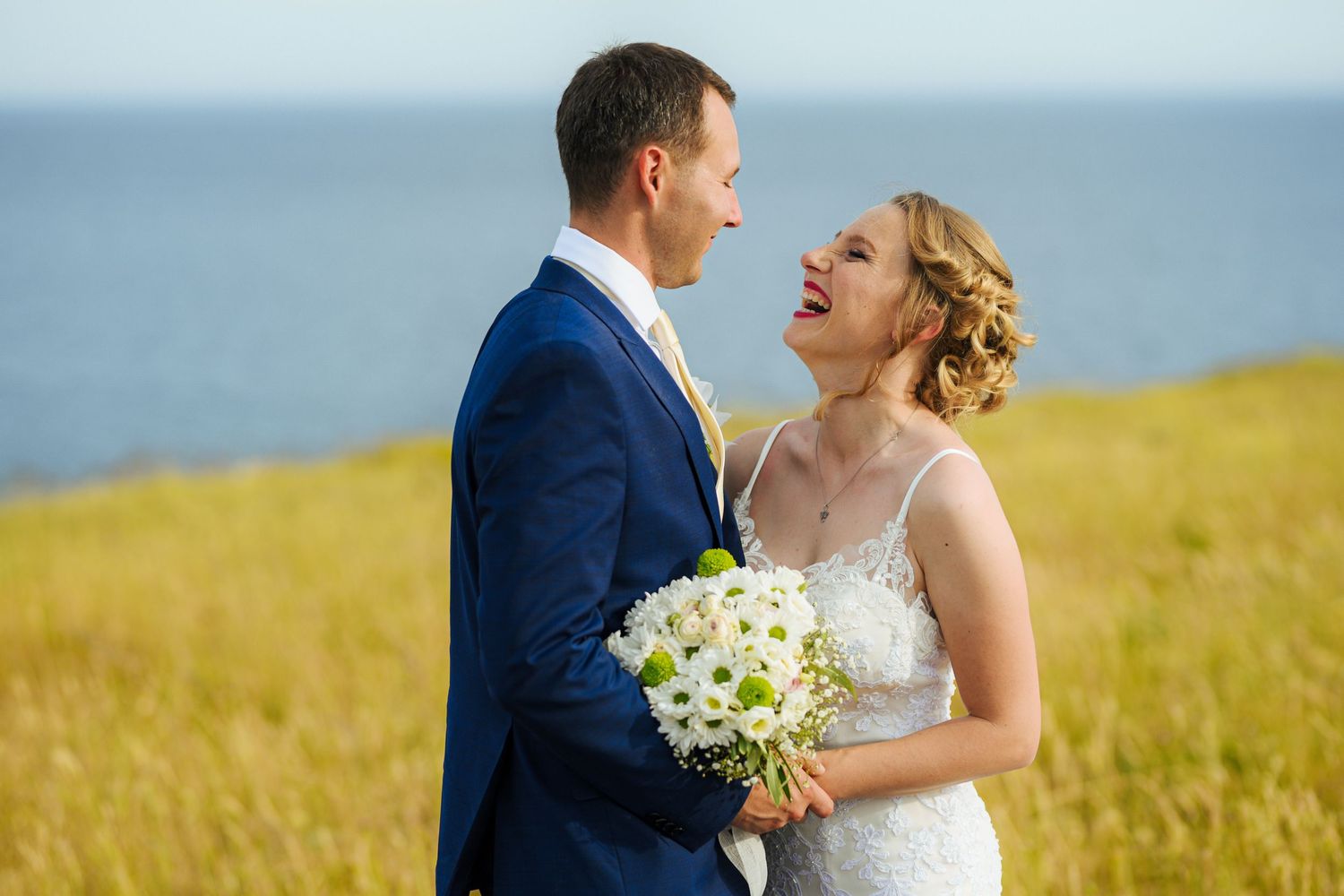 Laughing bride holding white bouquet stands close with groom in golden field by the sea.