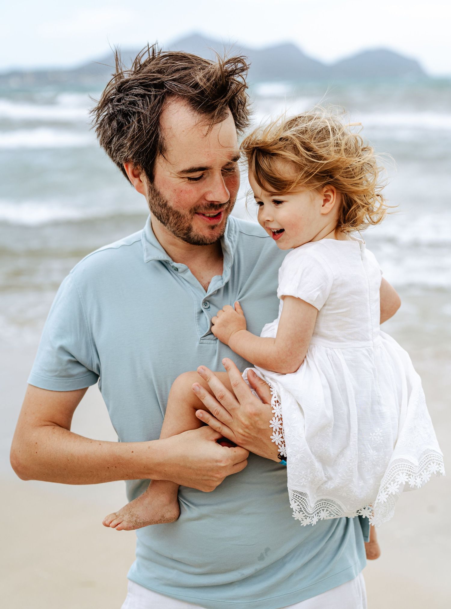 Dad smiling while holding laughing toddler daughter on sandy beach.