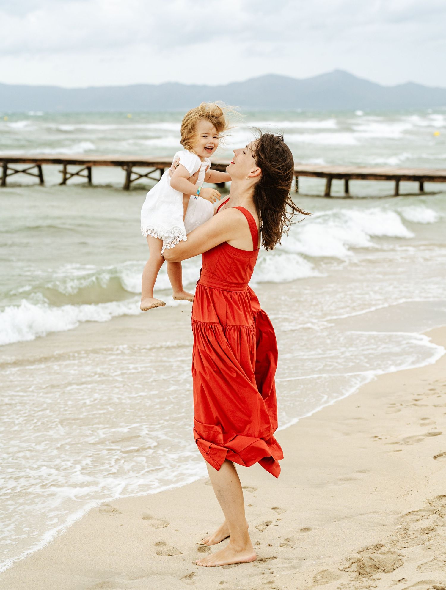 Mother in red dress holding laughing baby on windy beach with pier.
