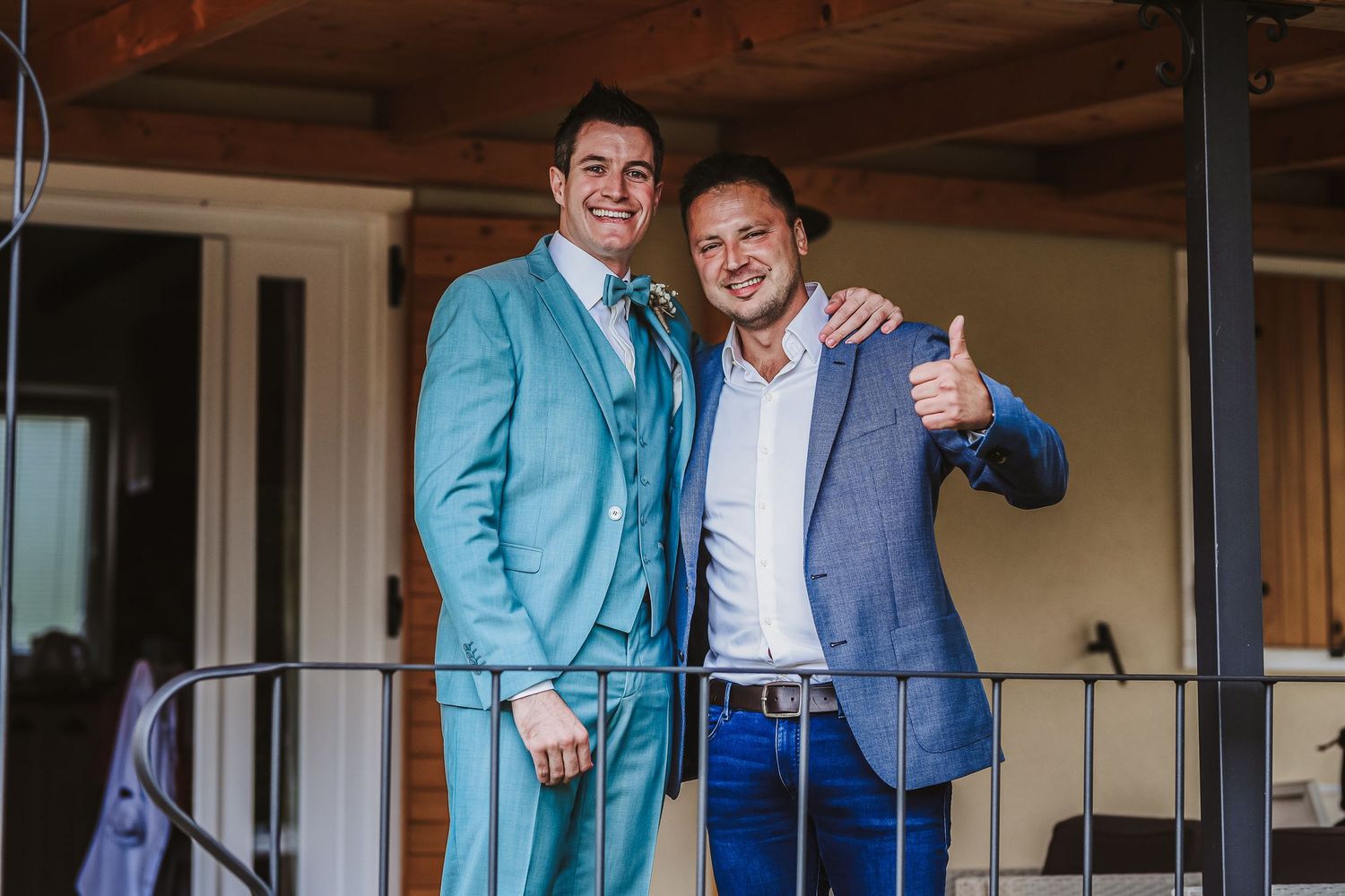 Two men in blue suits smiling and posing together on a balcony, one giving a thumbs up.