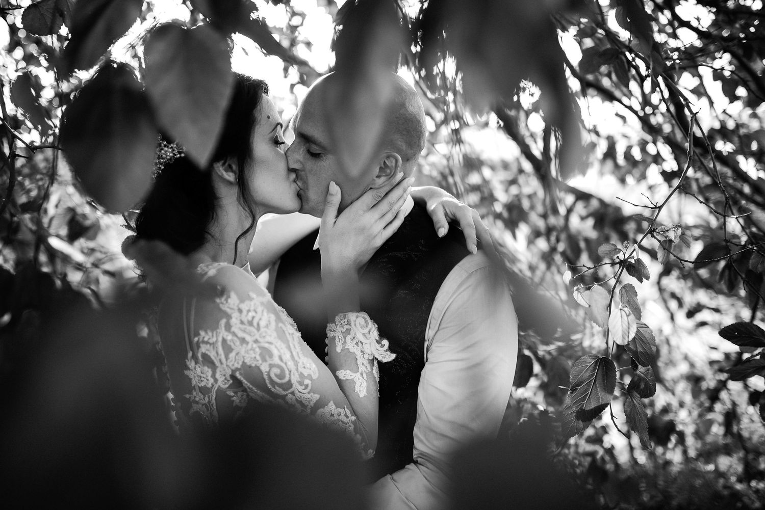 Bride and groom sharing a romantic kiss surrounded by lush green leaves in black and white.