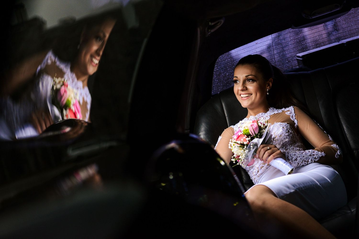 Smiling bride in white dress holds a floral bouquet while sitting in a dark luxury limousine.
