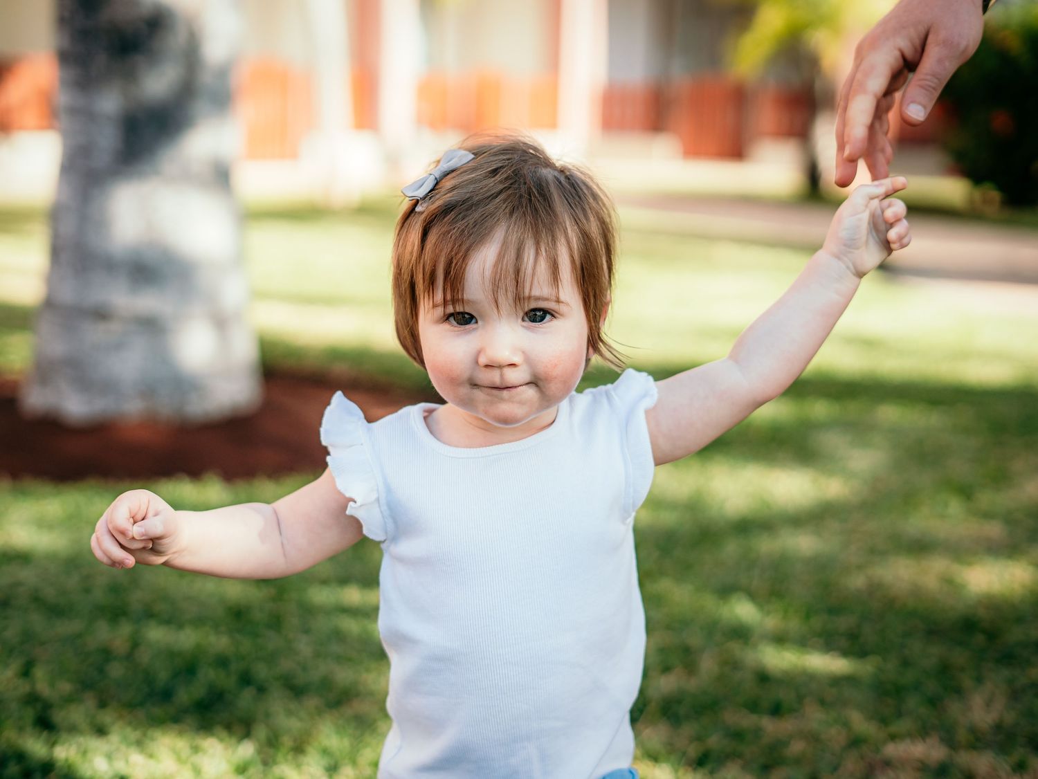 Happy baby girl in white top reaches up to hold an adult's hand while learning to walk on green grass.