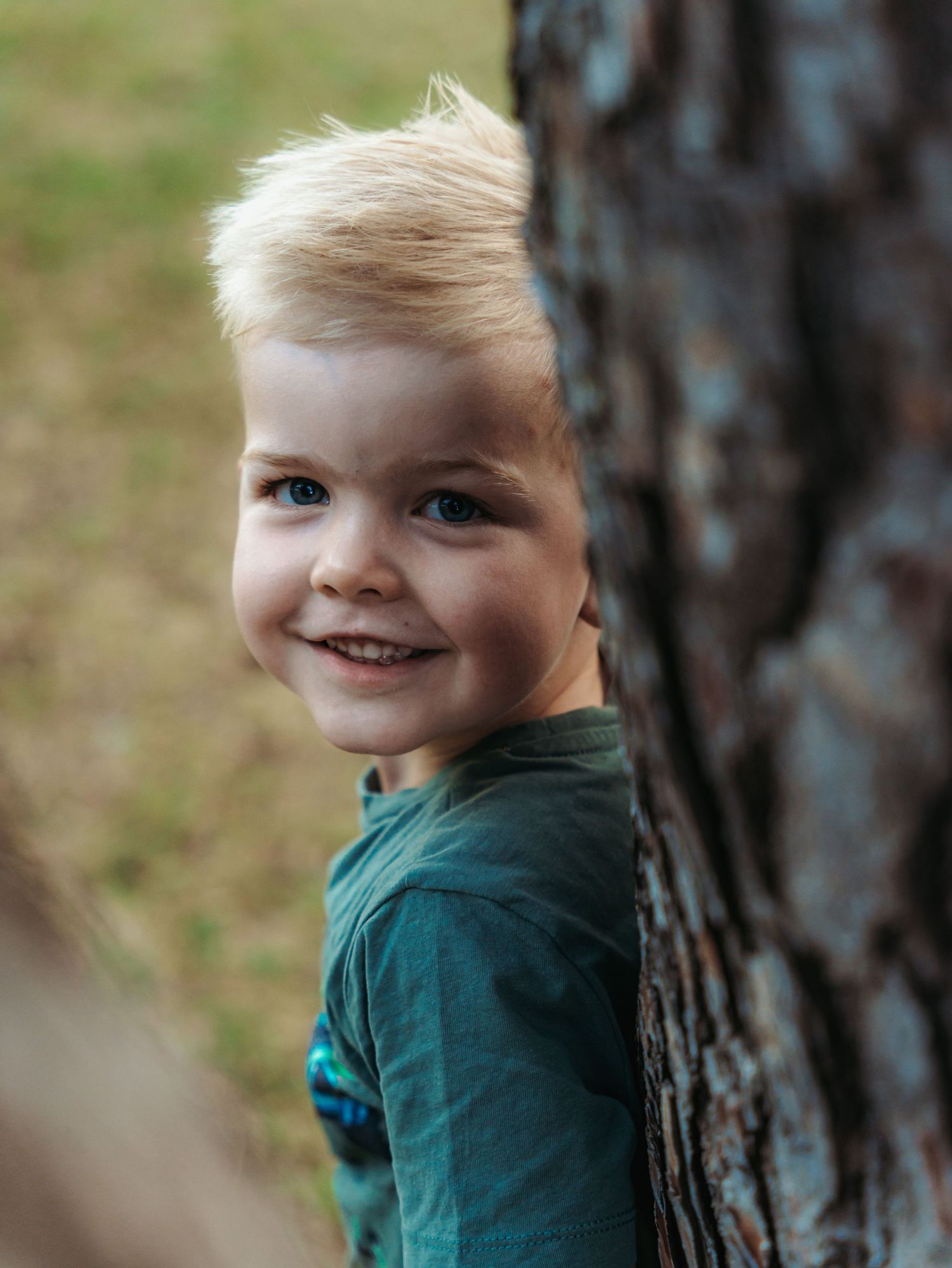 A smiling blonde boy in green shirt peeks playfully from behind a tree trunk outdoors.
