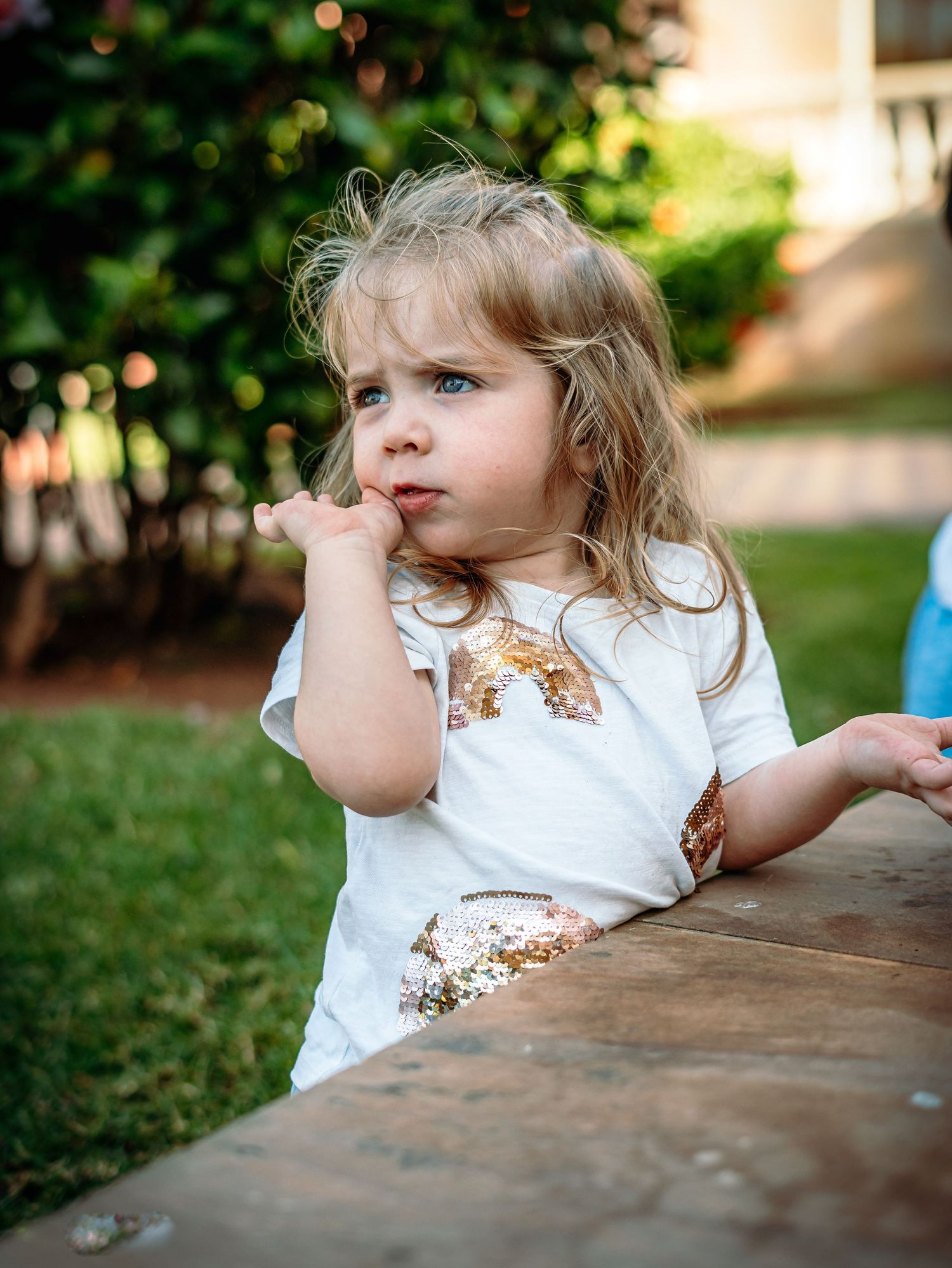 Adorable blonde toddler girl in sequin rainbow shirt resting chin on hand outdoors.