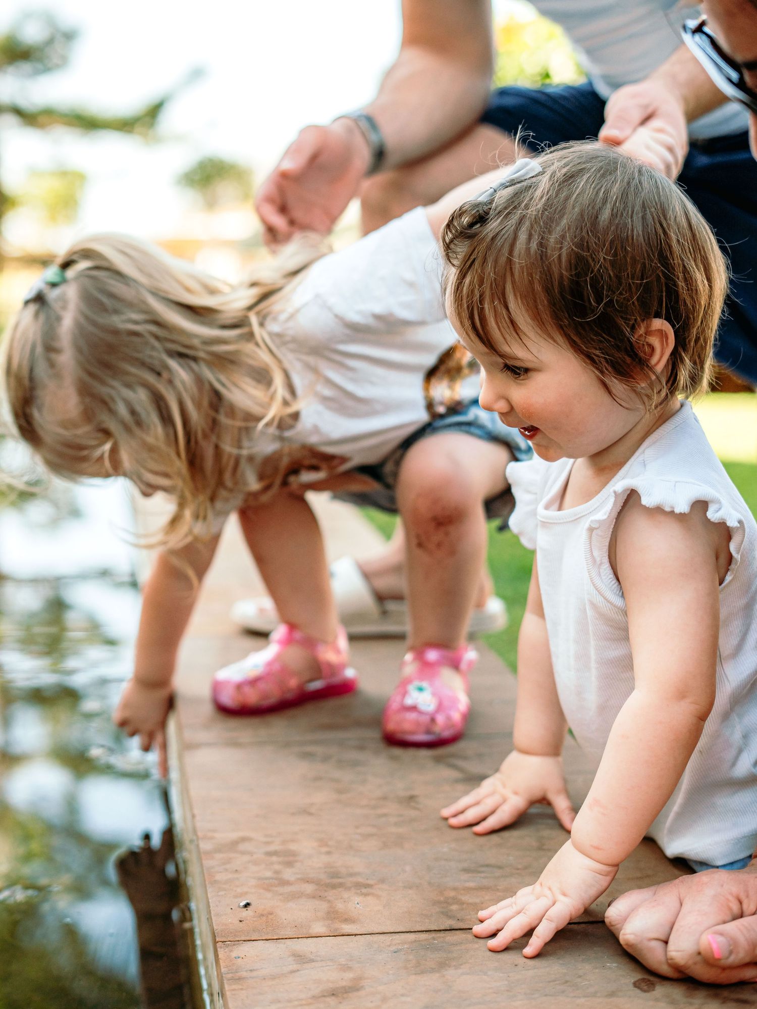 Two toddler girls in white tops crouch on a wooden ledge, exploring a garden pond together.