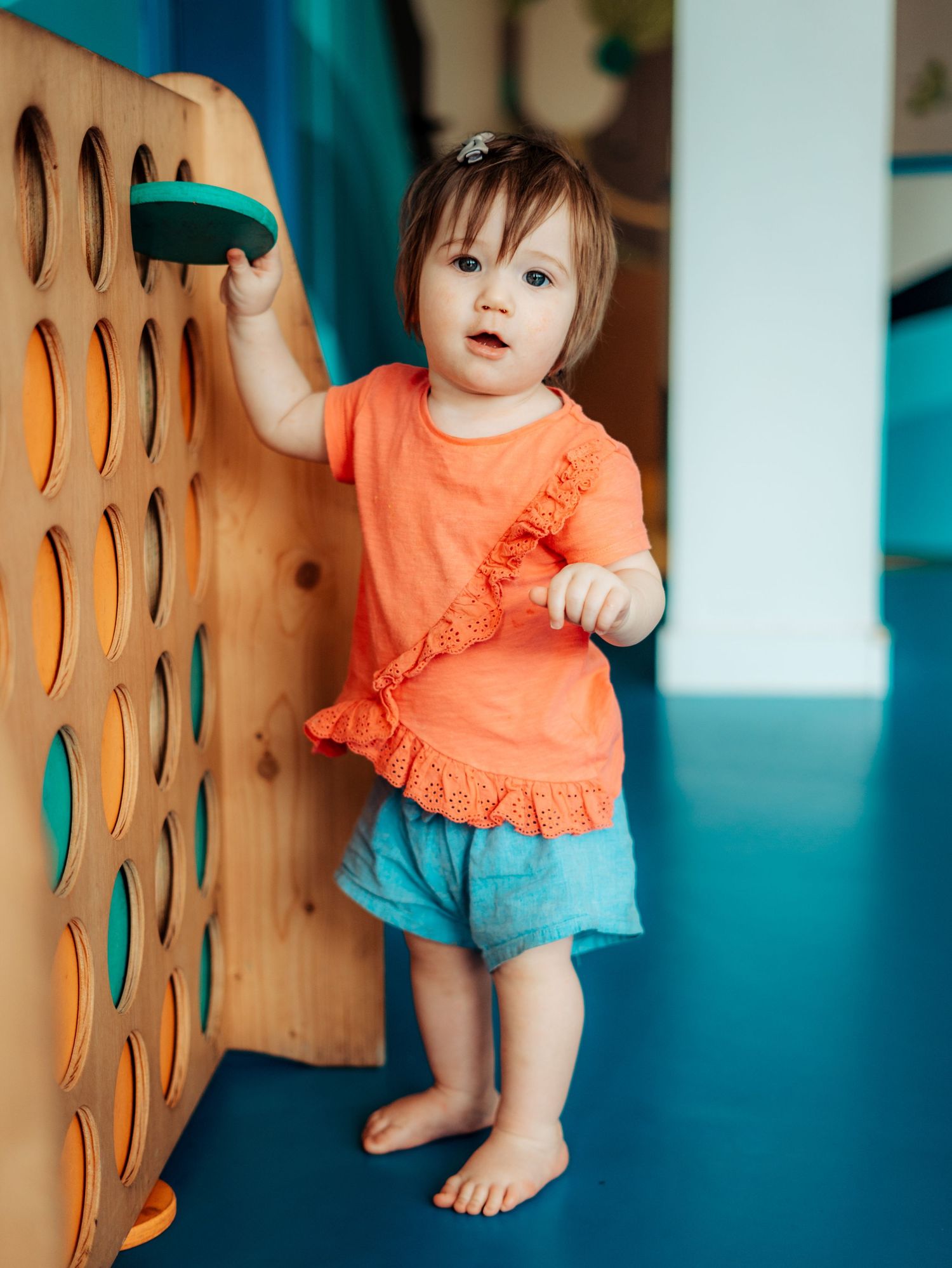 Barefoot toddler girl in orange top and blue shorts playing with teal toy at wooden frame.