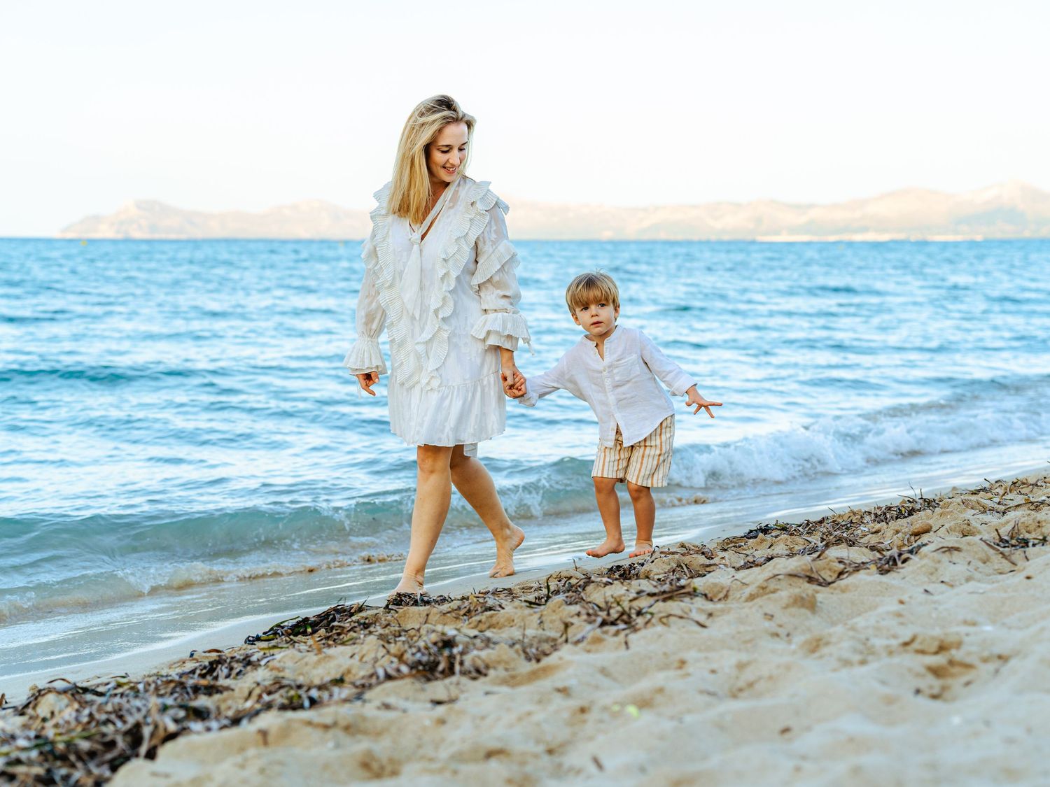 Mother and young son walking hand in hand along a sandy beach shore in matching white outfits.