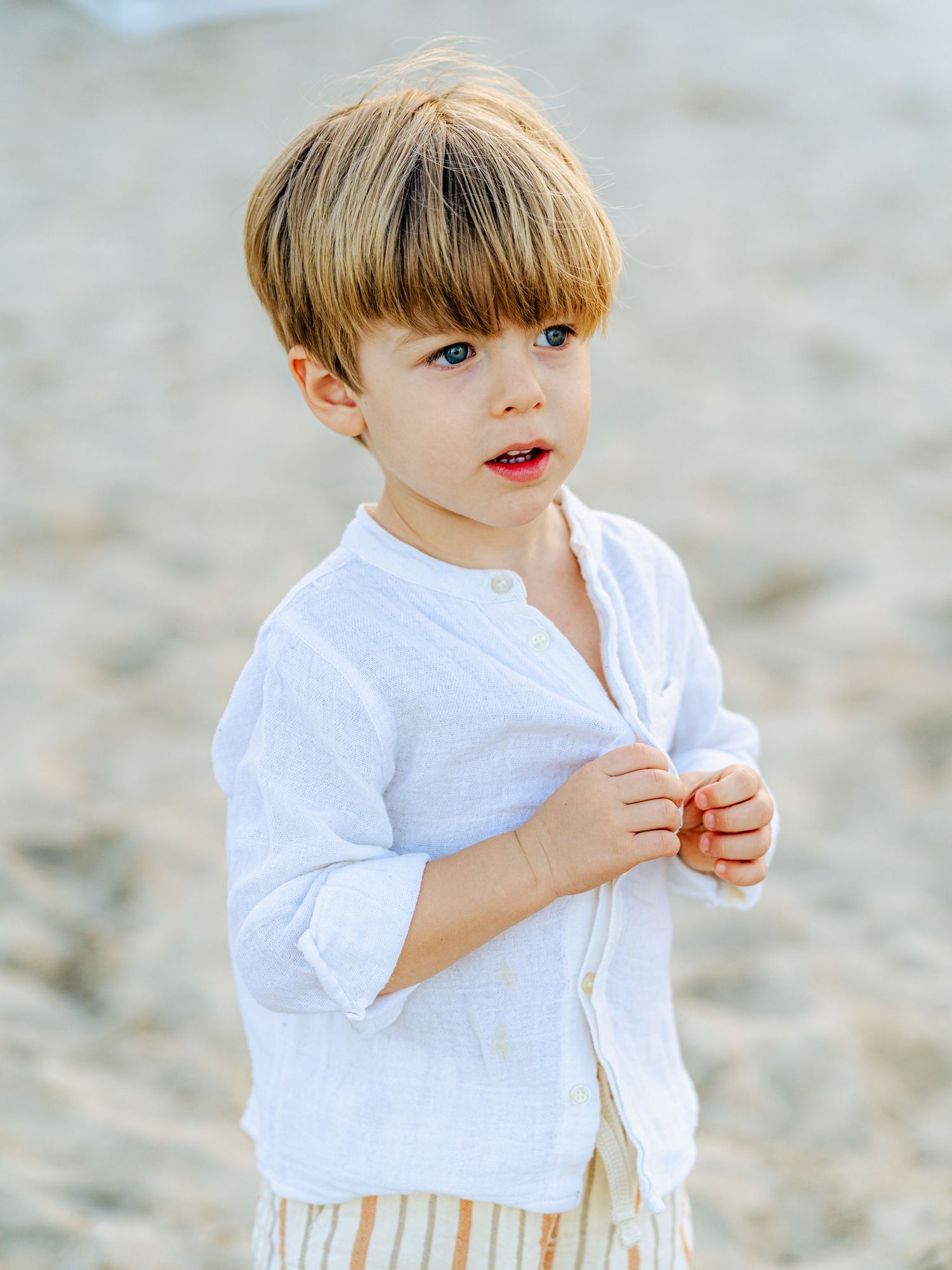 Young blonde boy in white linen shirt and striped shorts standing on sandy beach.