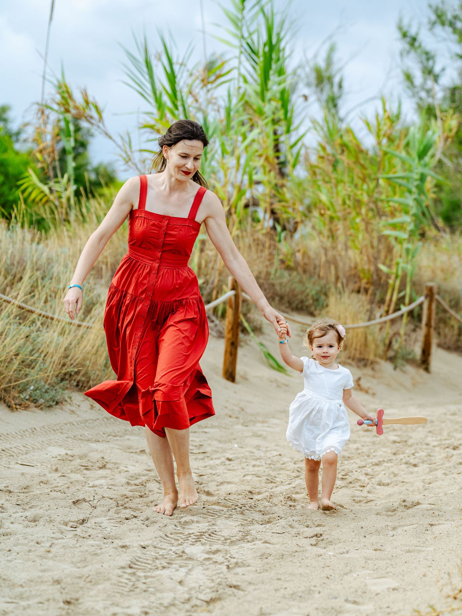 A mother in a red dress holds hands with her toddler in white on a sandy beach path.