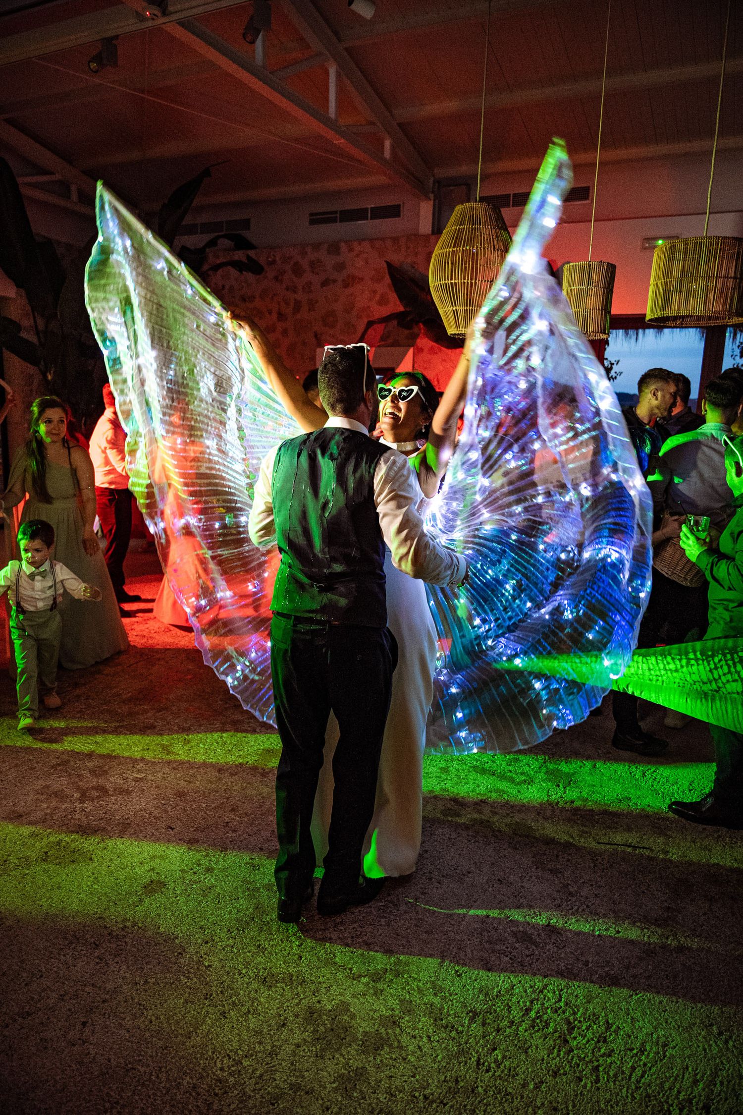 Wedding couple dances surrounded by a performer wearing spectacular illuminated LED wings at reception.