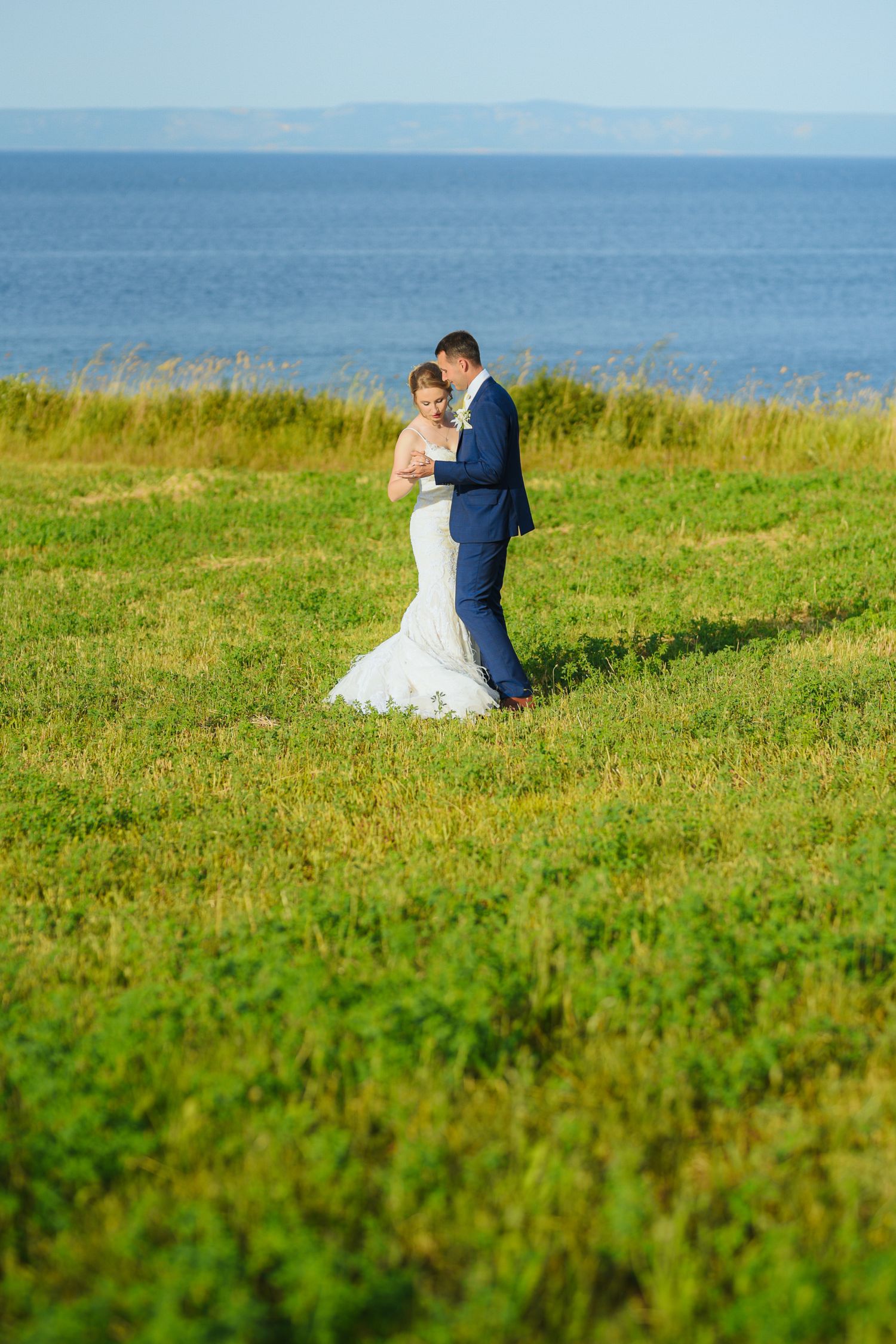 Bride and groom embrace in a lush green field with the blue ocean and distant mountains in the background.