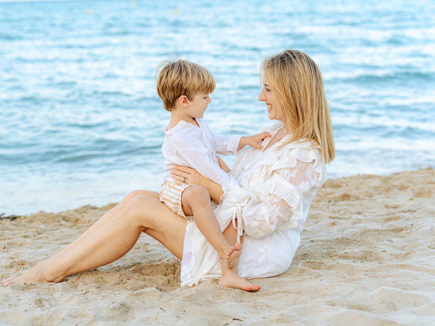 Happy mother and young son sitting together on sandy beach smiling at each other by the blue sea.