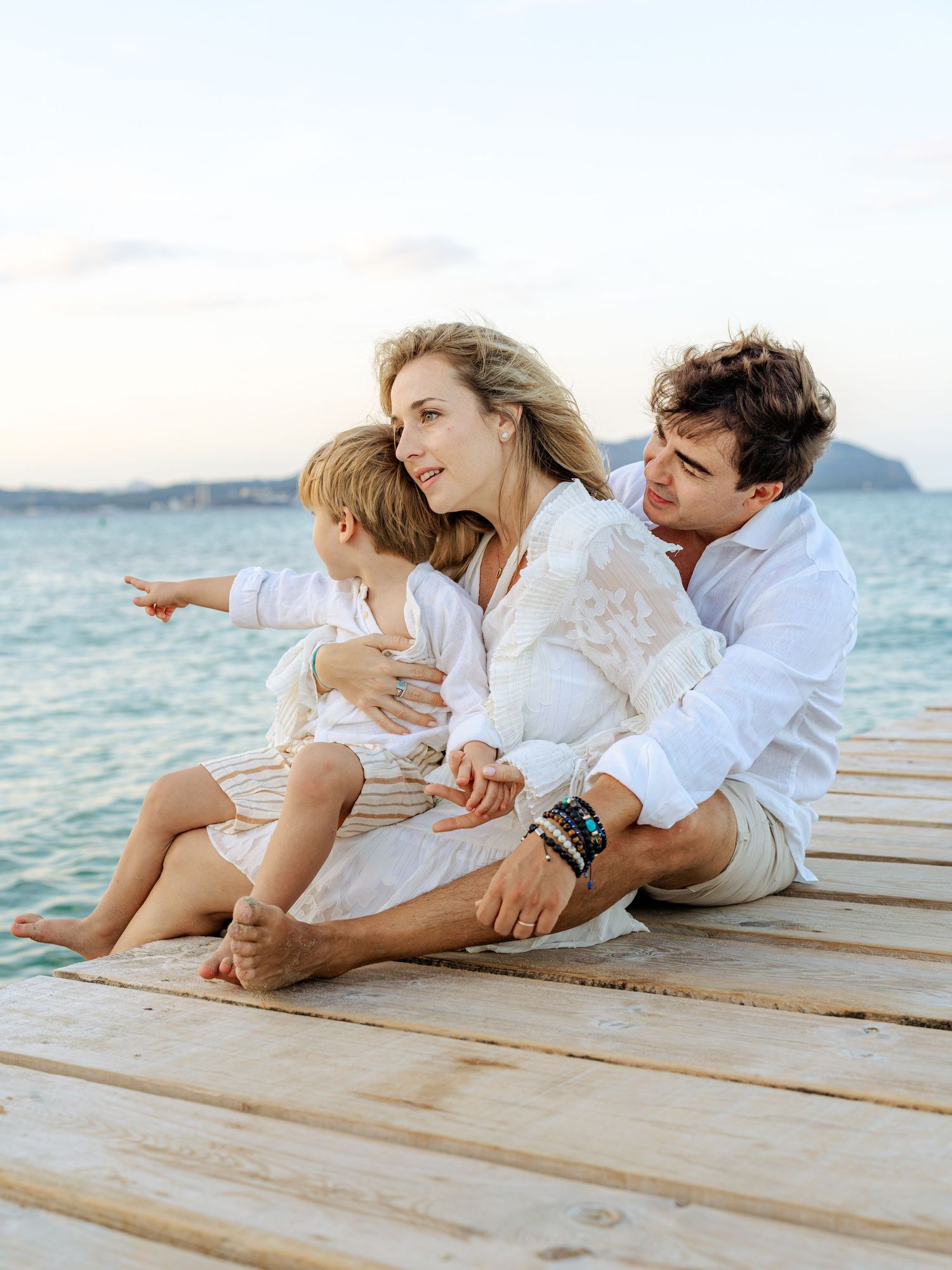 Family of three sitting on a dock at sunset, child pointing toward the calm blue ocean.