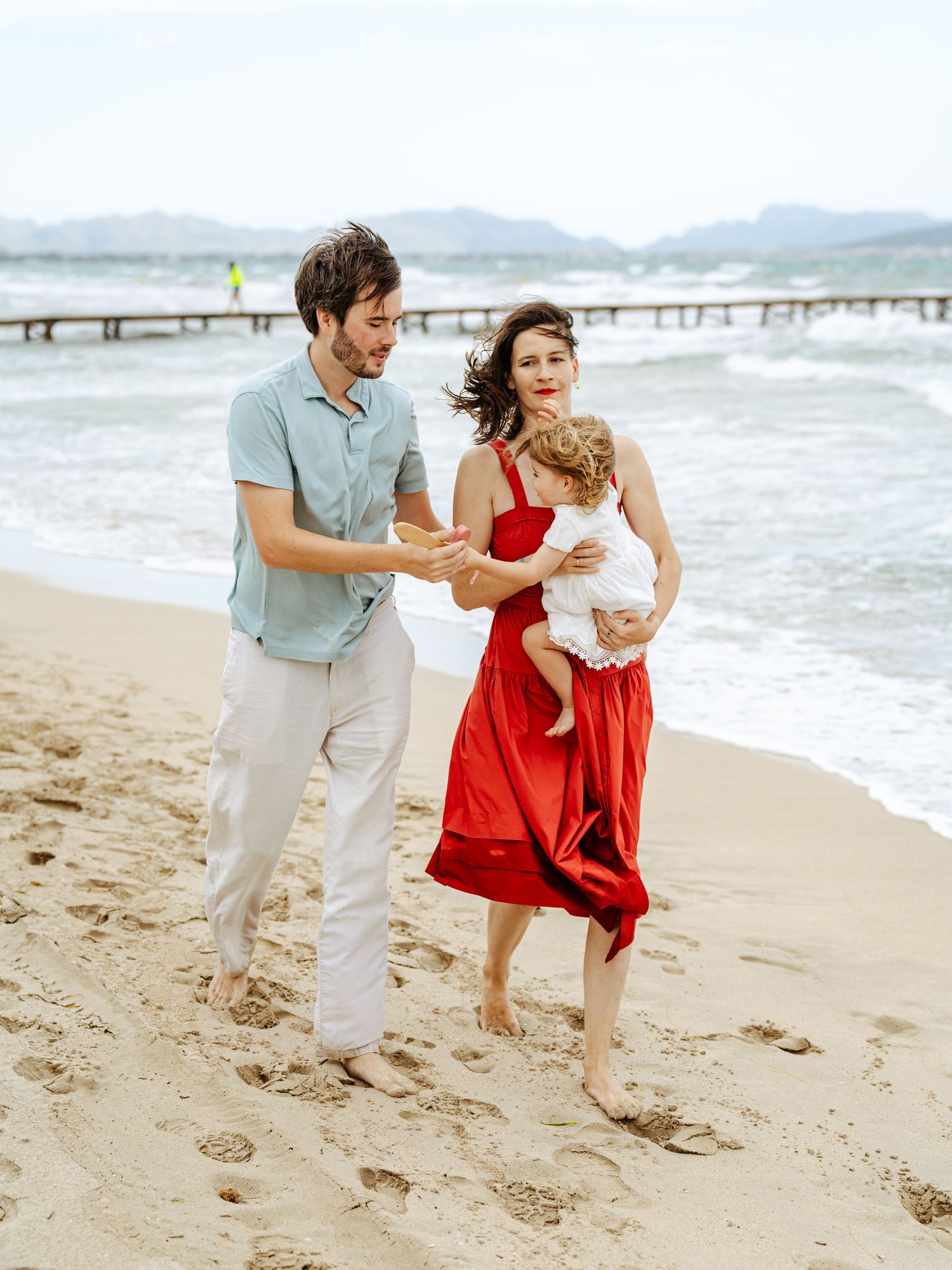 Happy family with father, mother in red dress, and baby enjoying a sunny beach day.