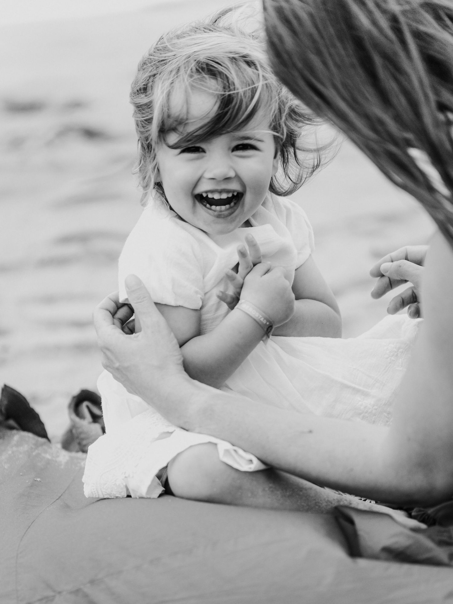 Black and white photo of a laughing toddler sitting on beach with mother nearby.