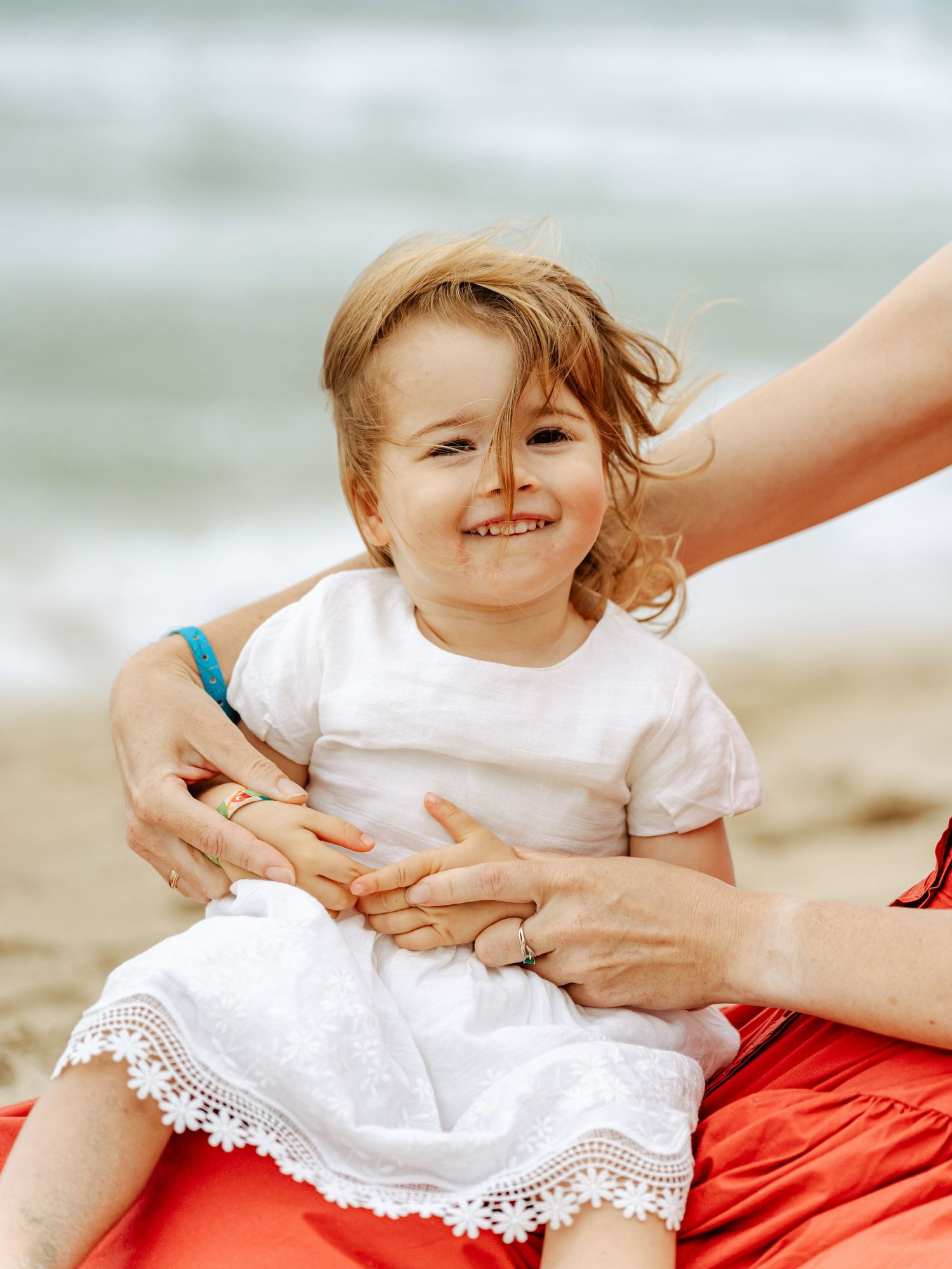 Smiling blonde toddler girl in white lace dress held by mother on red blanket at beach.
