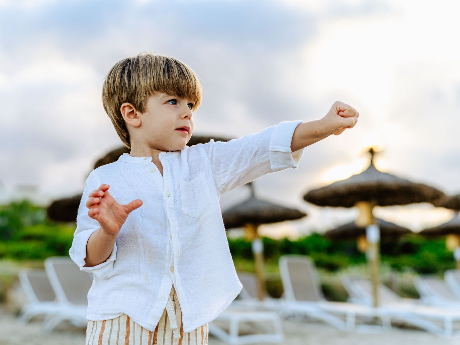 Young boy in white shirt practicing karate moves on a beach with thatched umbrellas in background.