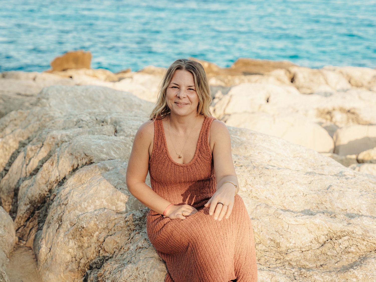 Smiling blonde woman in rust knit dress sitting on rocky shore with blue ocean behind her.