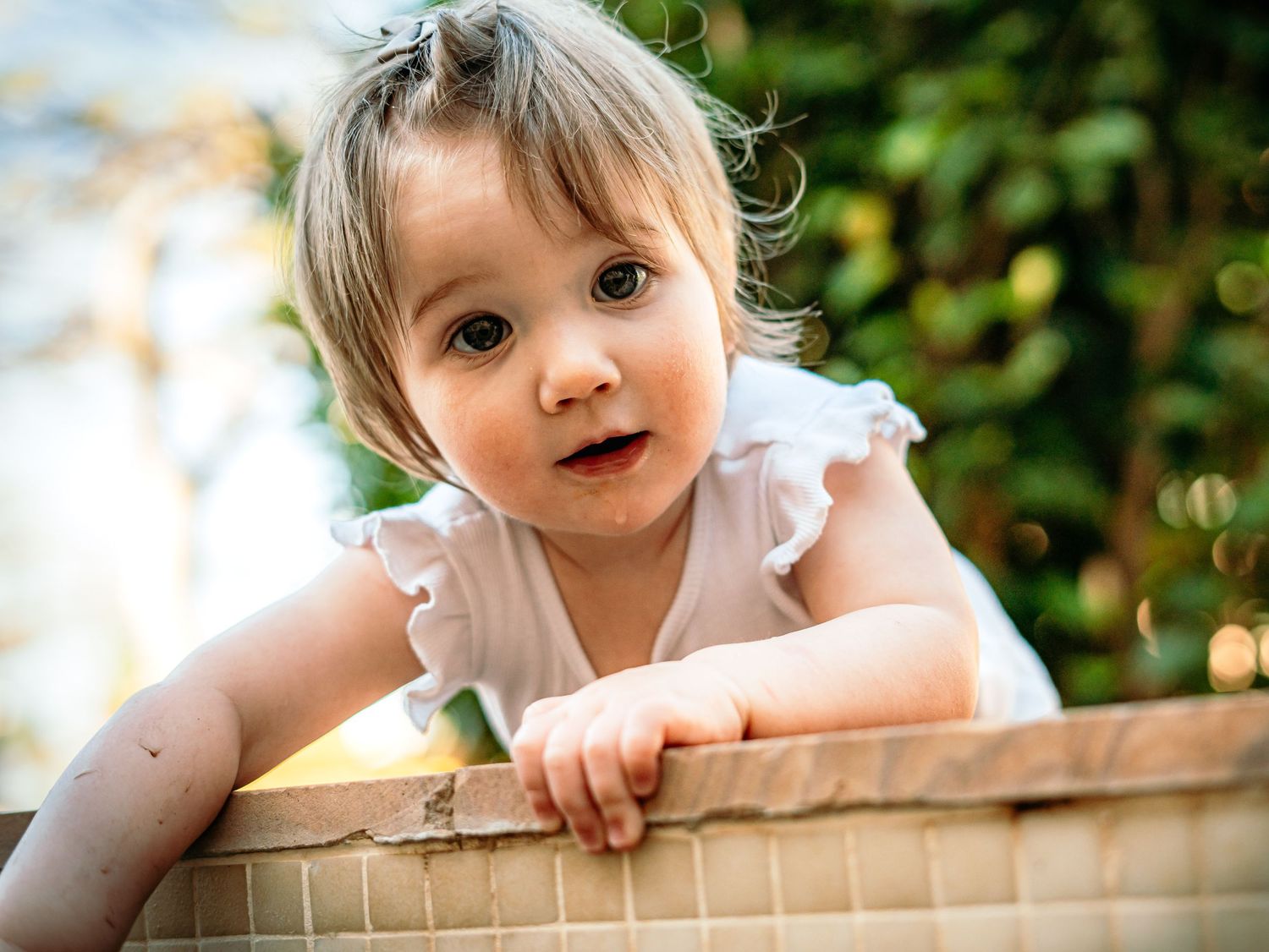 Curious toddler girl in white dress peering over a wooden ledge outdoors.