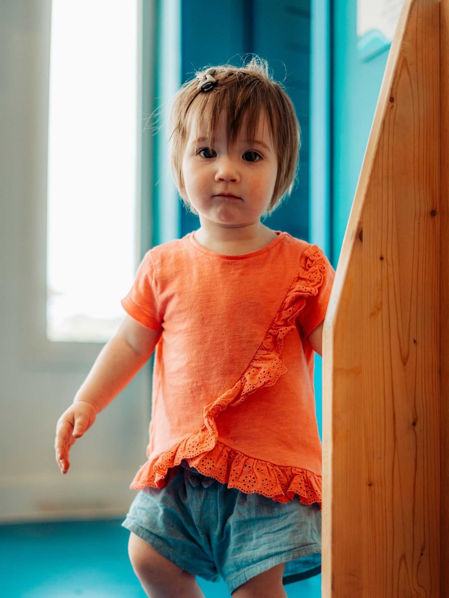 Toddler girl in orange ruffled top and denim shorts standing beside a wooden play structure indoors.