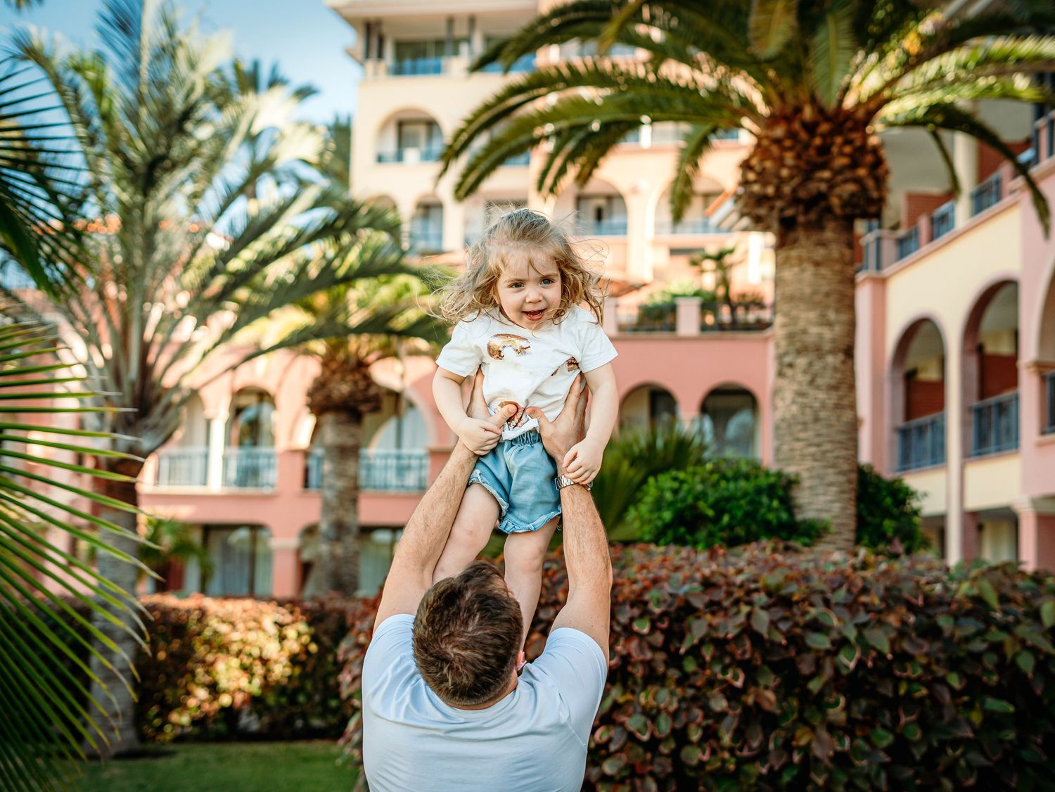 Father lifting happy toddler girl high in the air near tropical palm trees at resort.