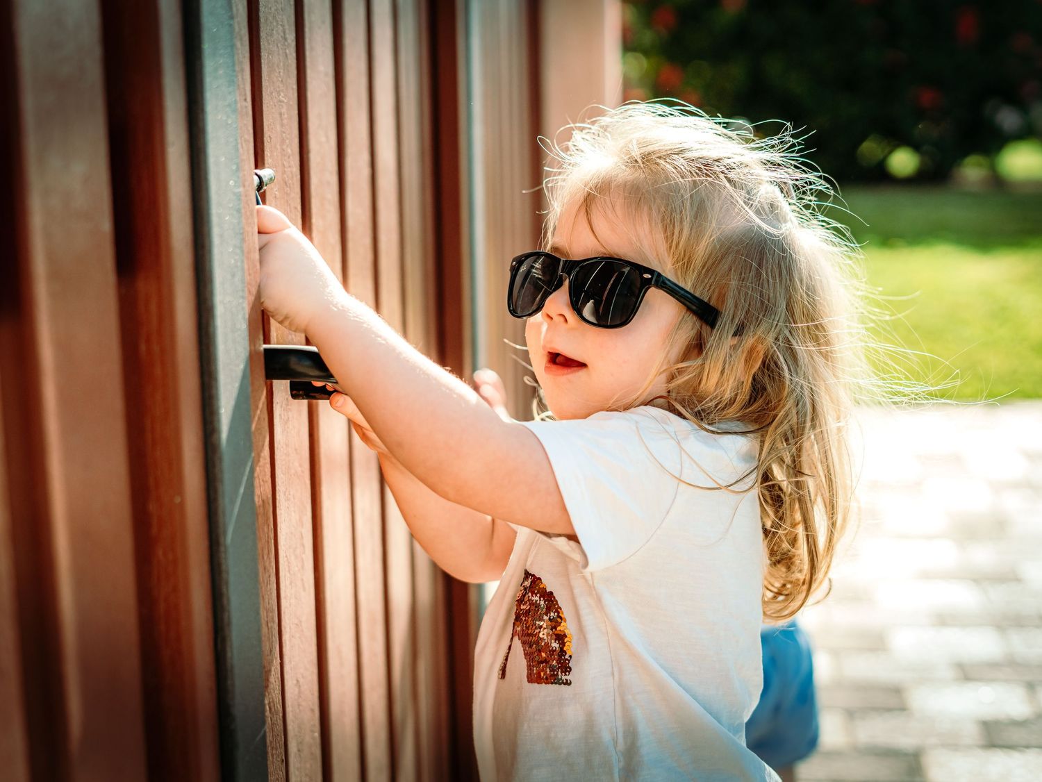 Cute toddler girl with long blonde hair wearing oversized black sunglasses reaching for a wooden fence gate.