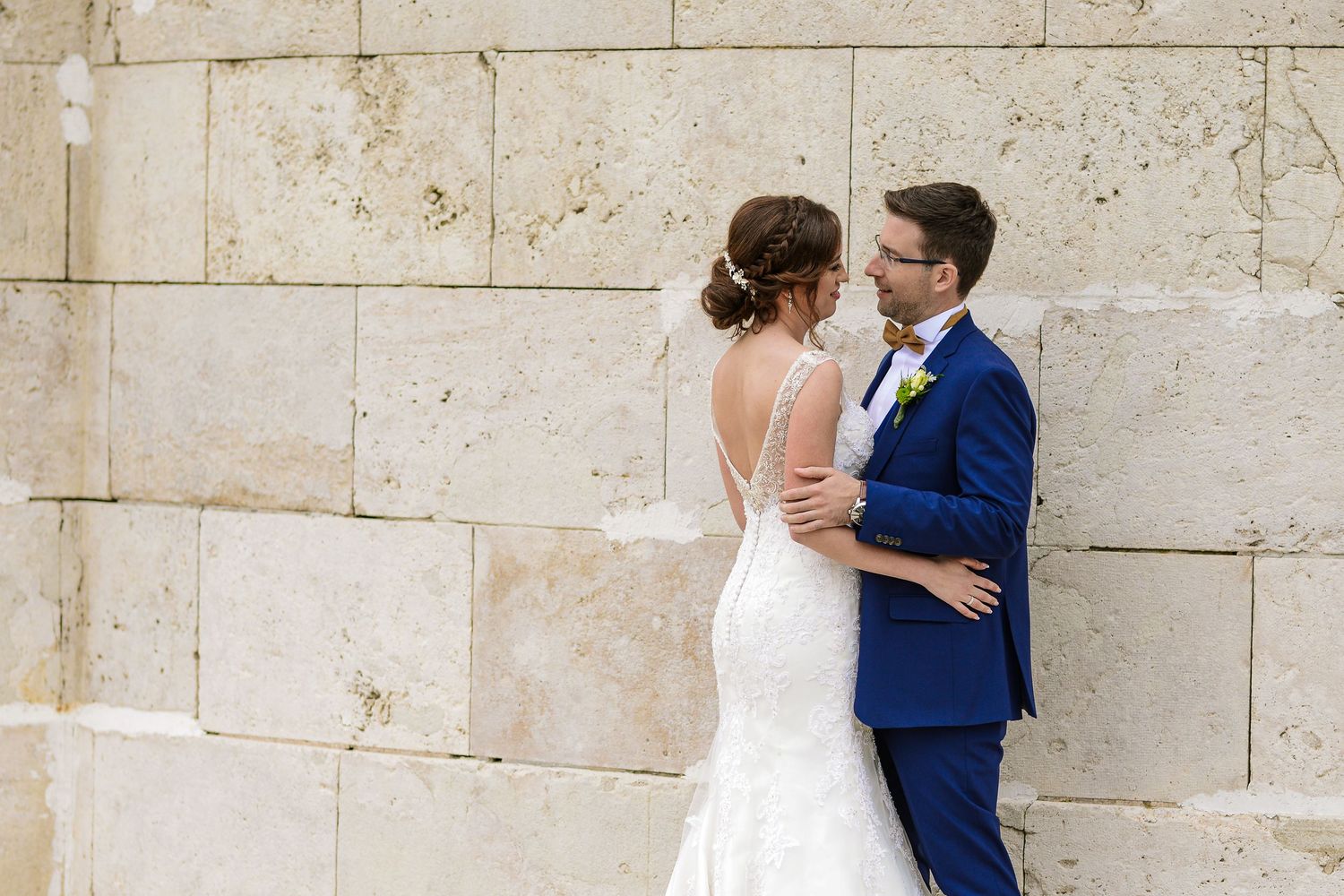 Bride and groom embrace romantically against a stone wall, she in a backless lace gown, he in a navy blue suit.