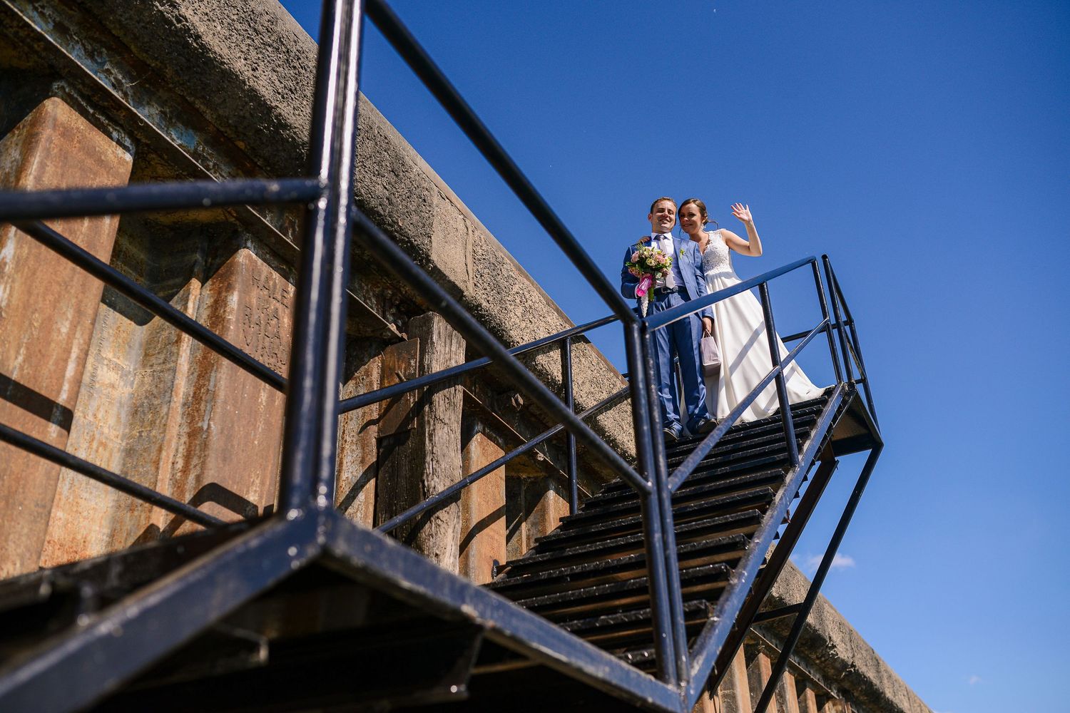 Bride and groom standing on metal stairs against a historic stone wall under a bright blue sky.