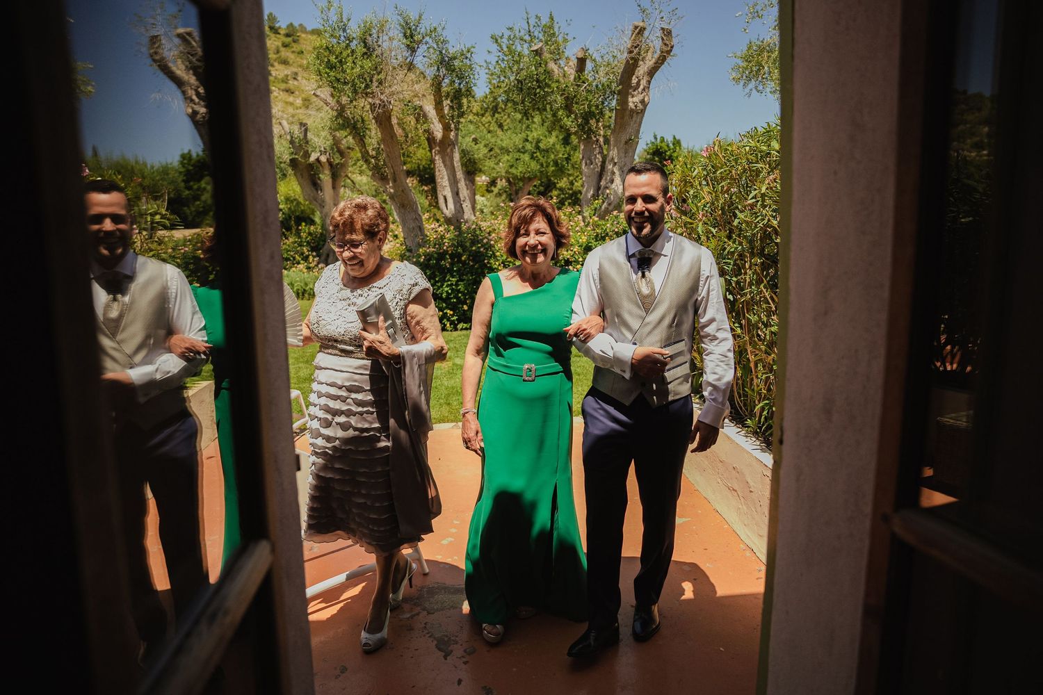 Three wedding guests in formal attire walking through an open door into a sunny garden.