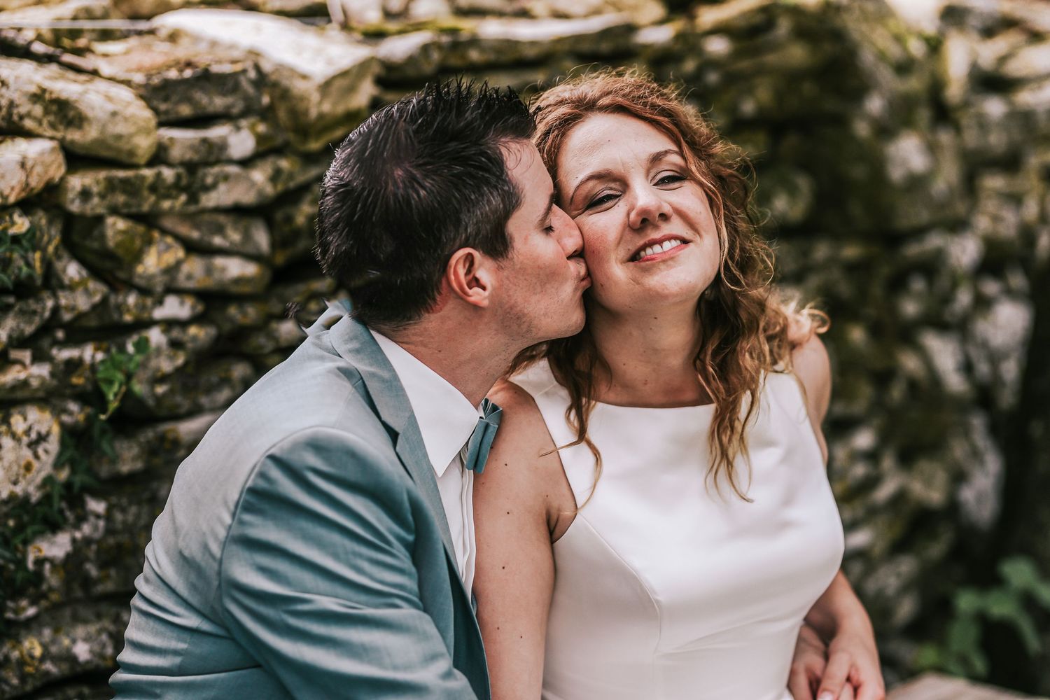 Groom kissing smiling bride on the cheek beside a rustic stone wall outdoors.