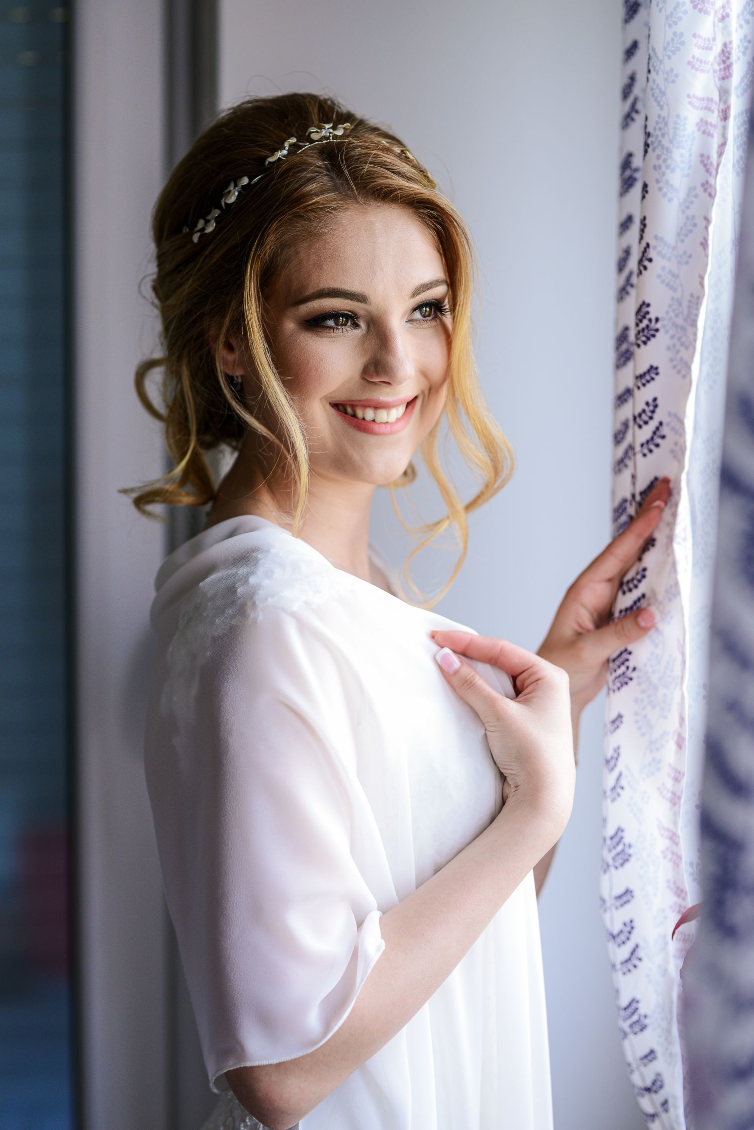 Smiling blonde bride in white robe with floral hair accessory poses by a curtained window.