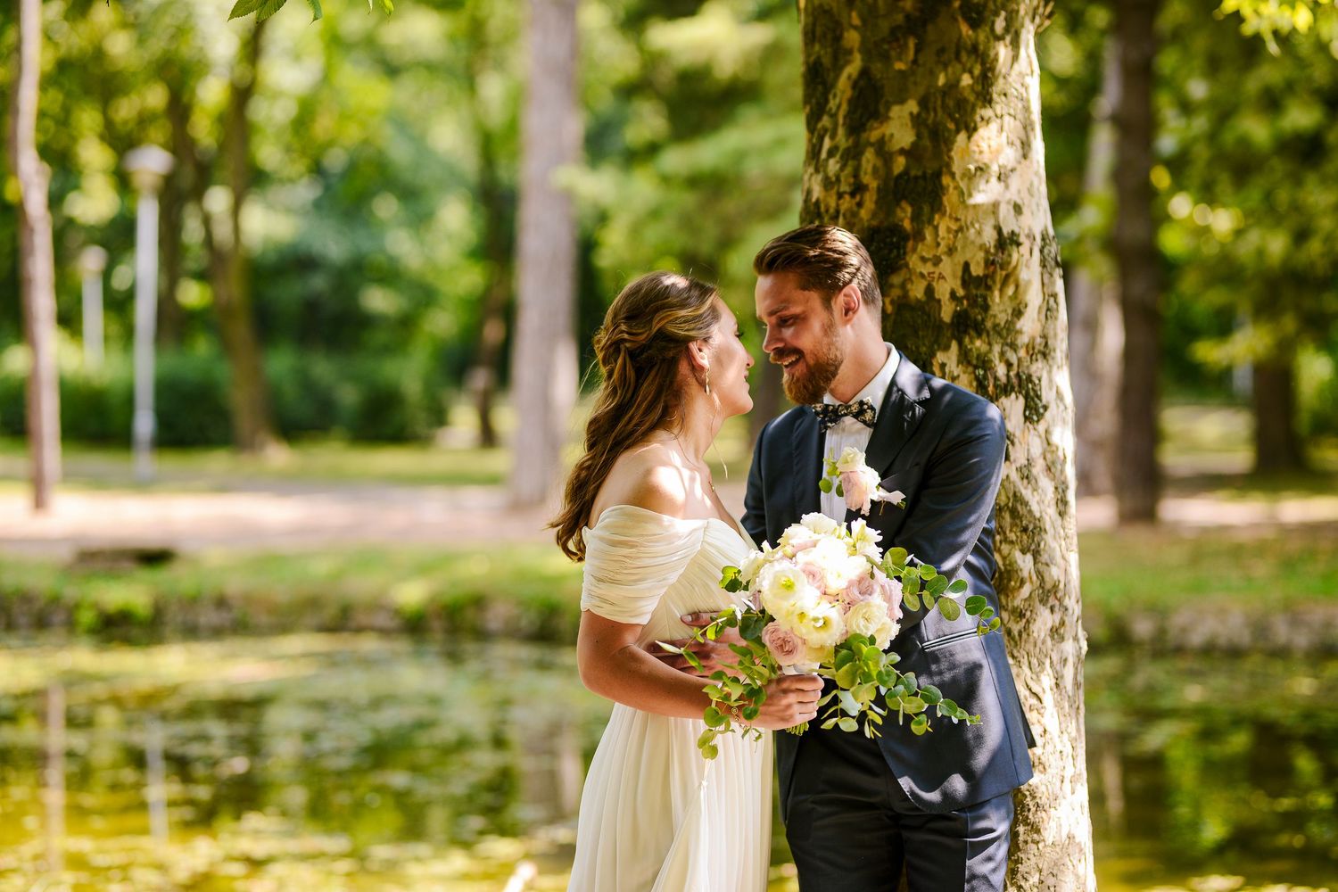 Smiling bride and groom share a tender moment by a pond, surrounded by sunlit green trees.