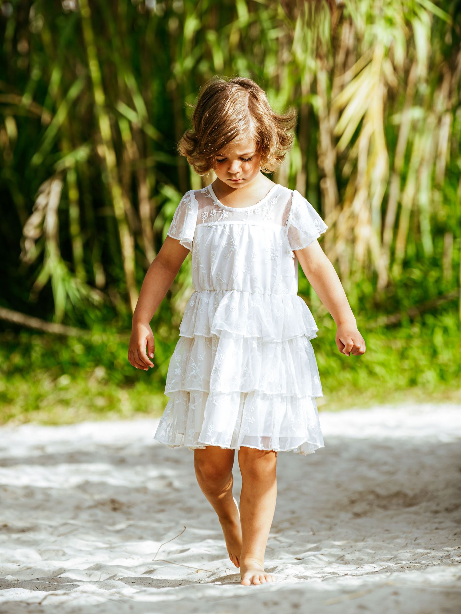 Young girl in white ruffled dress walks barefoot on sandy beach surrounded by greenery.