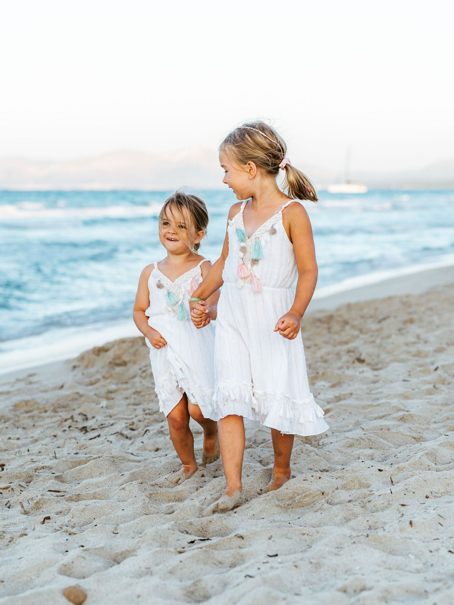 Two young sisters in white dresses walking hand-in-hand along a sandy beach at sunset.