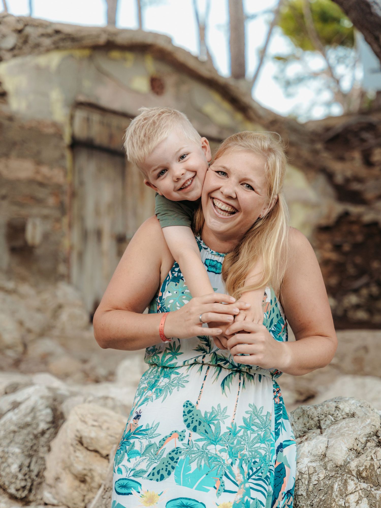 Smiling blonde mother in floral dress hugging her young blonde son outdoors near rustic stone wall.