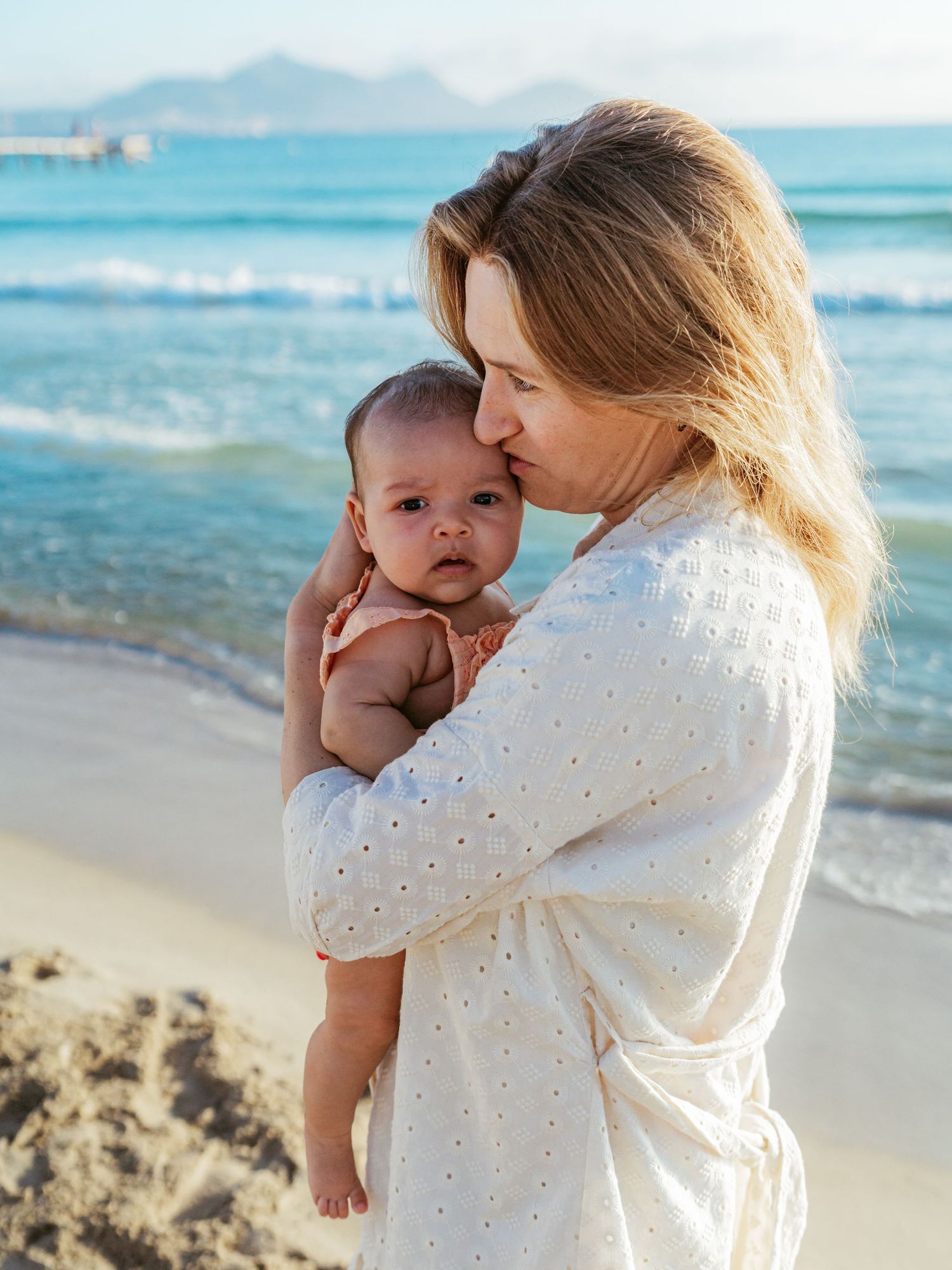 Blonde mother tenderly kissing her newborn baby on a sunny beach by the sea.