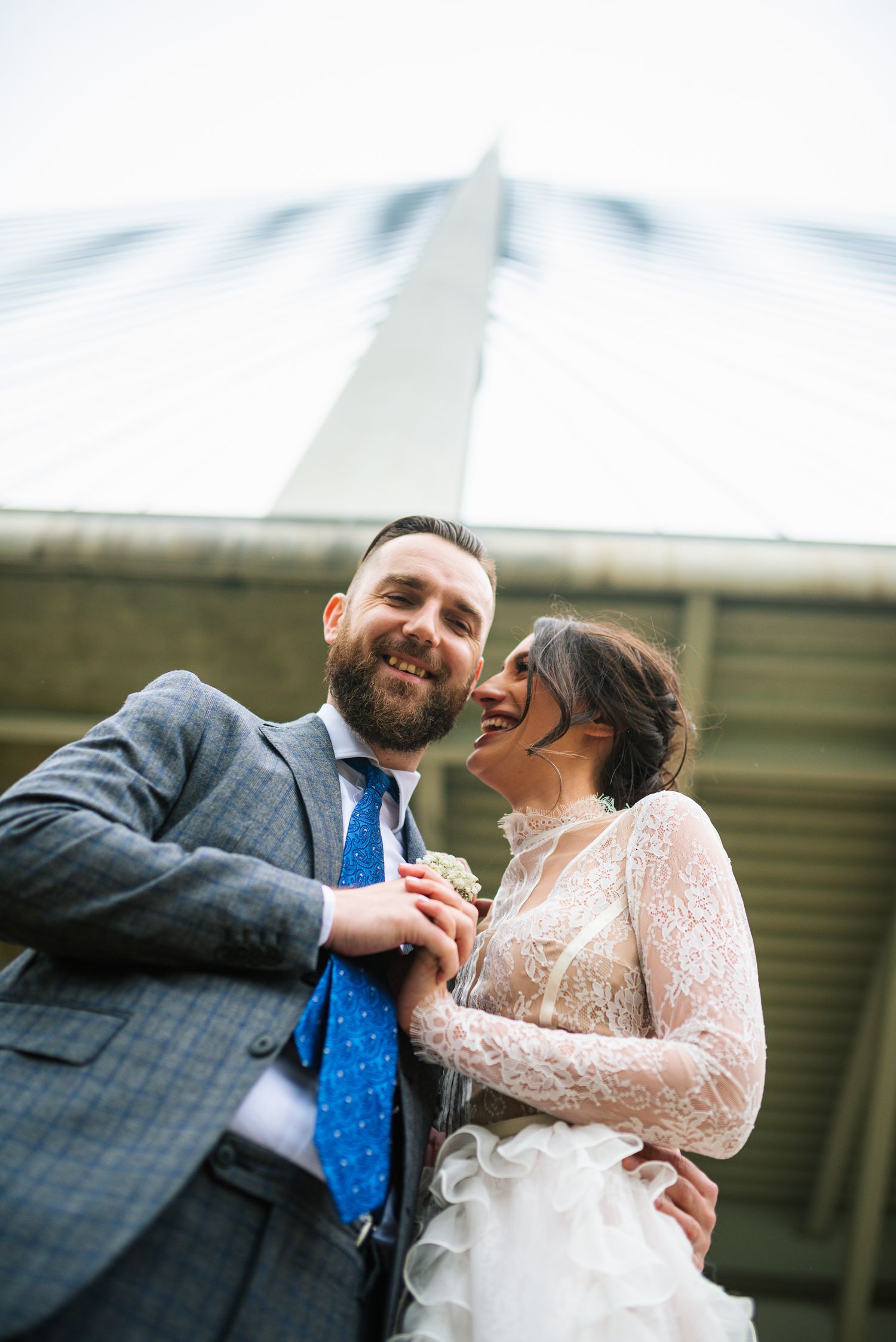 Low angle view of laughing bride and groom holding hands beneath tall bridge structure on wedding day.