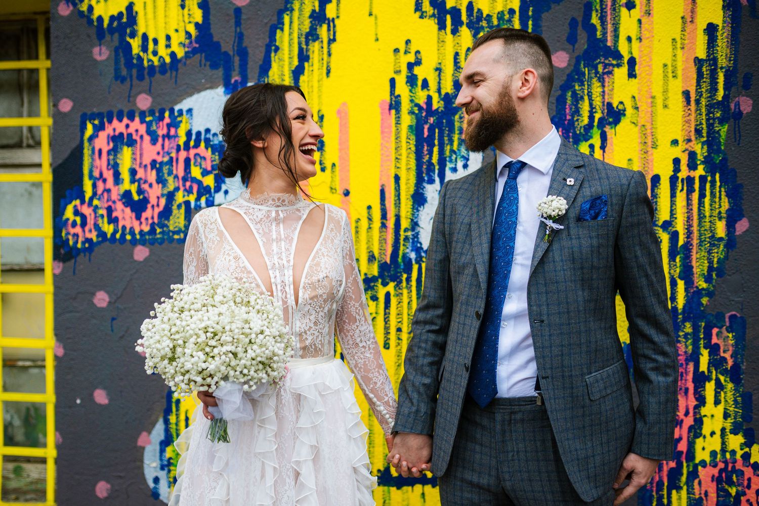 Laughing bride and groom holding hands in front of vibrant yellow and blue abstract graffiti wall.
