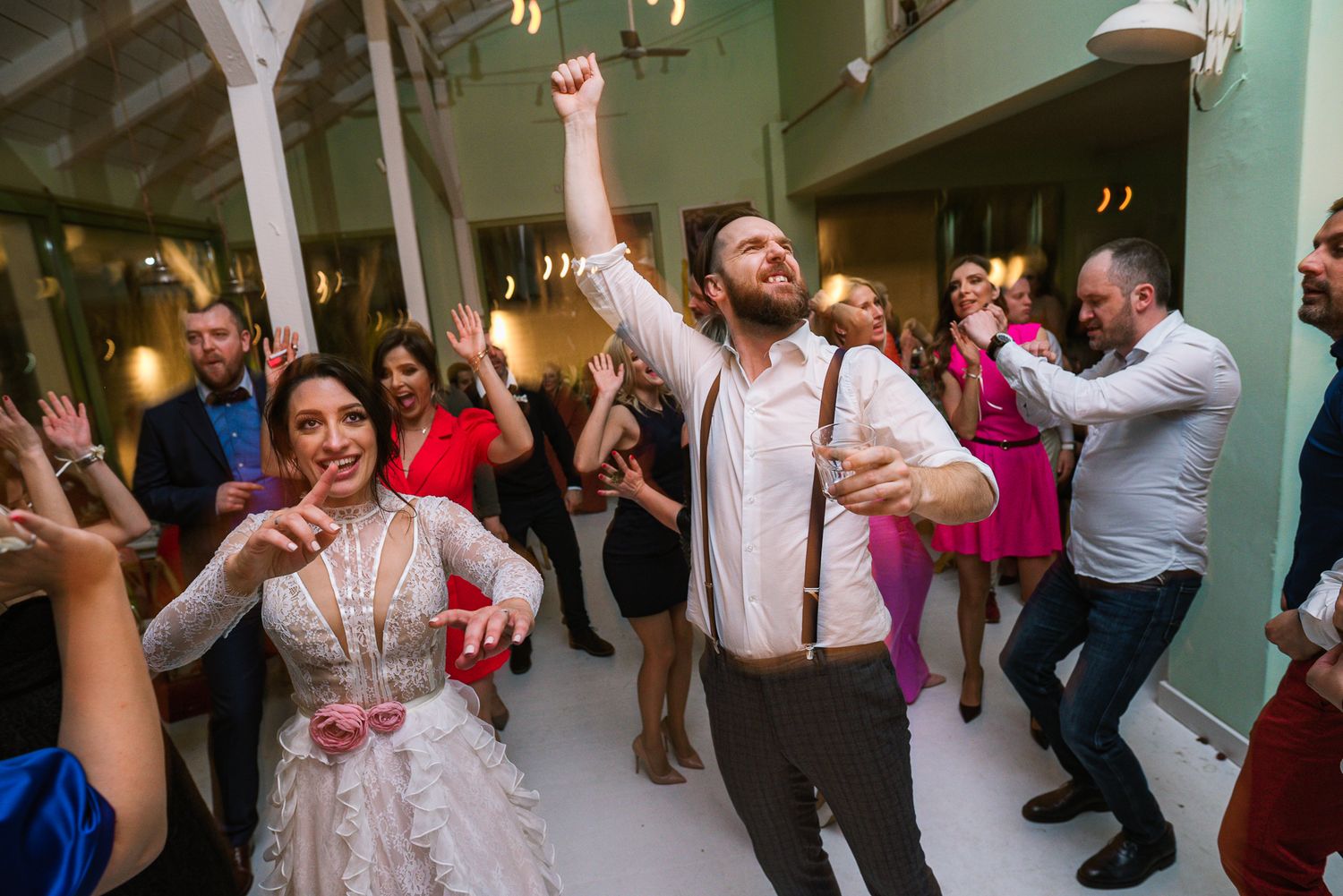 Bride and bearded groom dance energetically with guests at lively wedding reception.