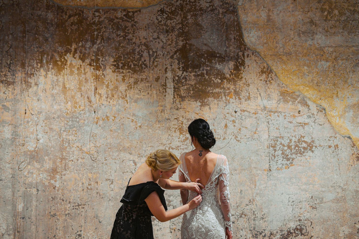 Blonde bridesmaid fastening buttons on back of brides fitted lace wedding gown.